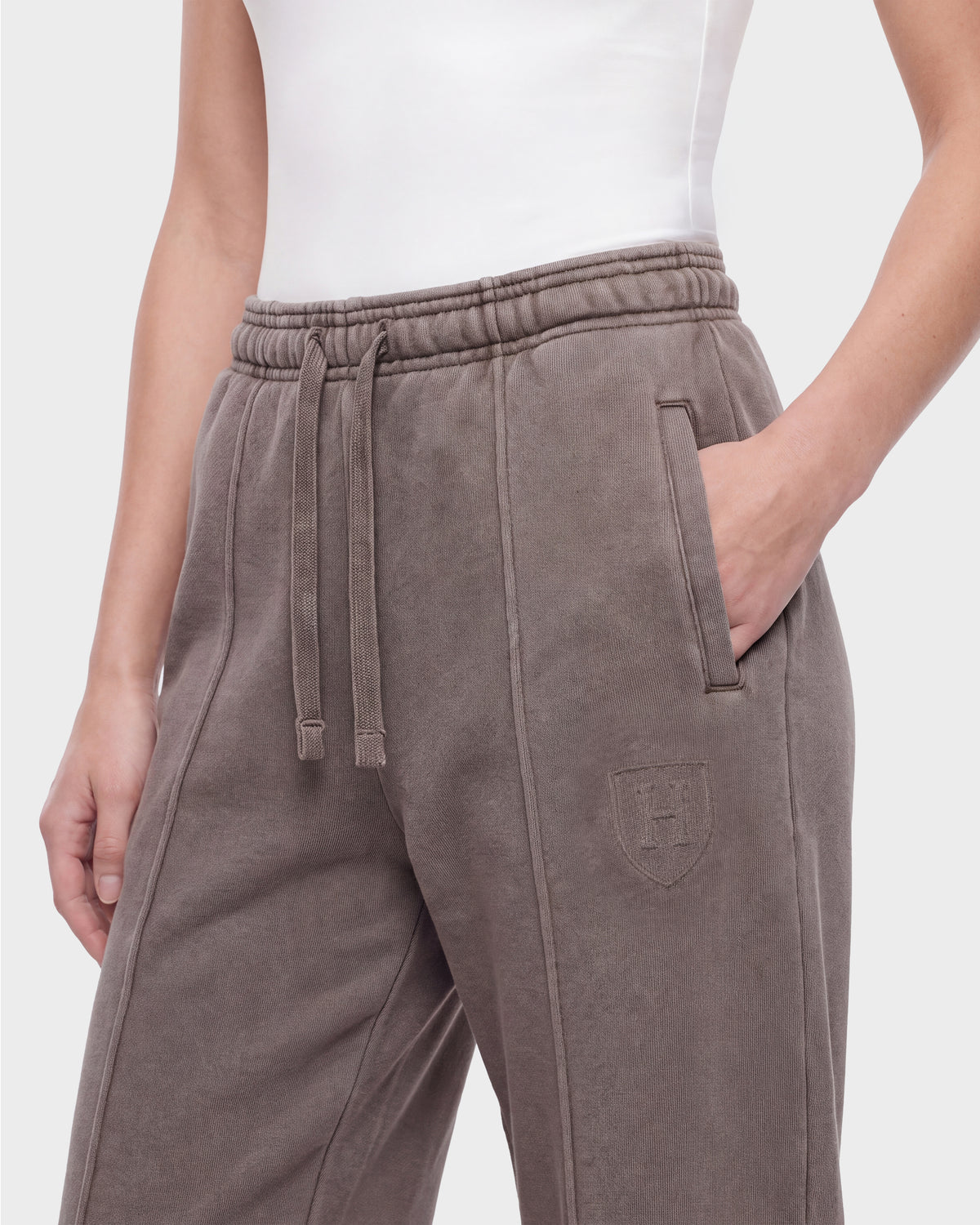 A person is wearing GAELLE HV - H526902 Taupe heavyweight jersey jogger sweatpants by Rudsak, featuring a drawstring waist and a crest logo on the thigh, paired with a white sleeveless top. Their left hand is in the pocket.