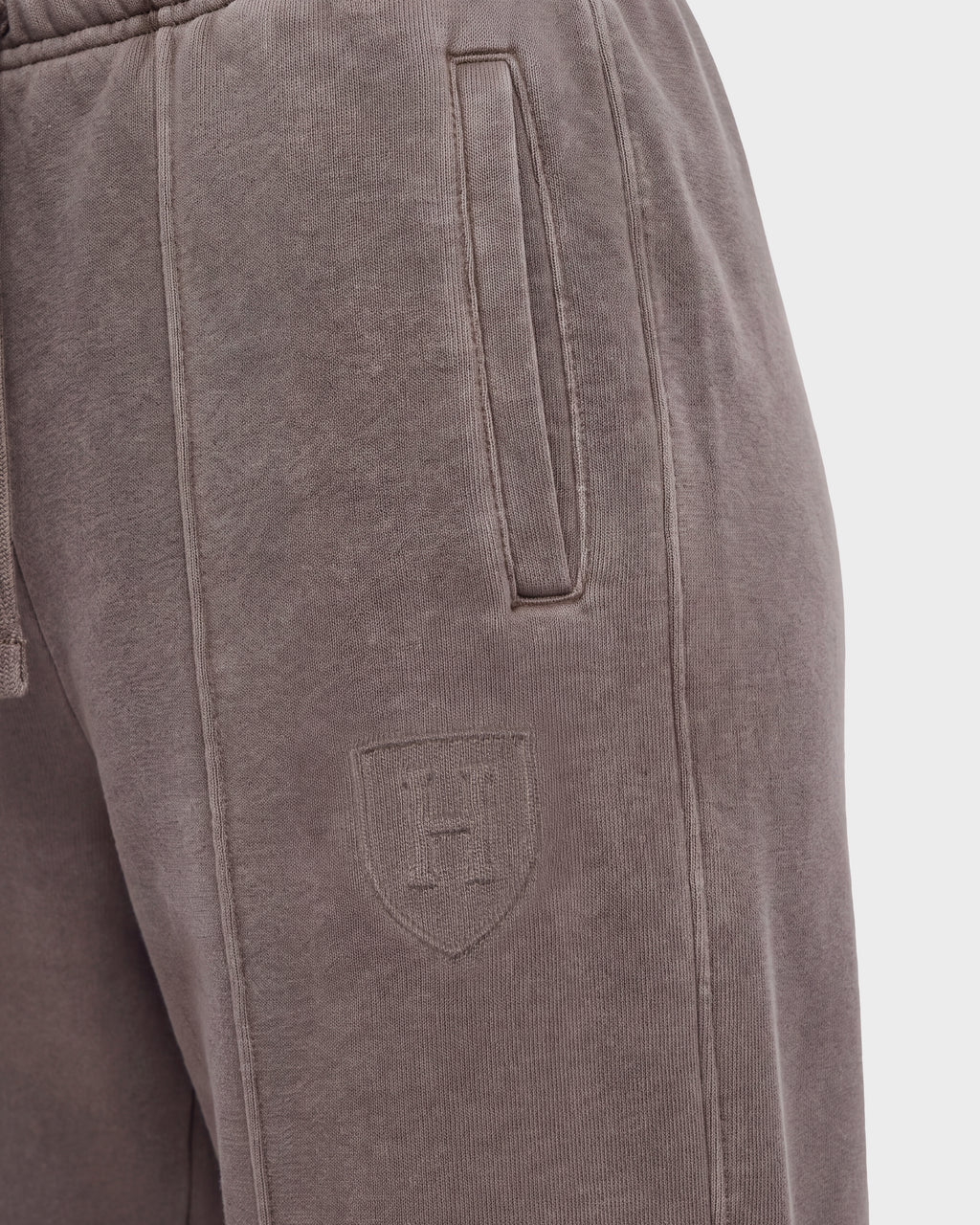 Close-up of the GAELLE HV - H526902 Taupe sweatpants by Rudsak, designed in a heavyweight jersey jogger style with a vertical side pocket and an embossed shield logo featuring the letter H on the upper thigh. The fabric appears soft and slightly textured, evoking the timeless appeal of classic Harvard apparel.