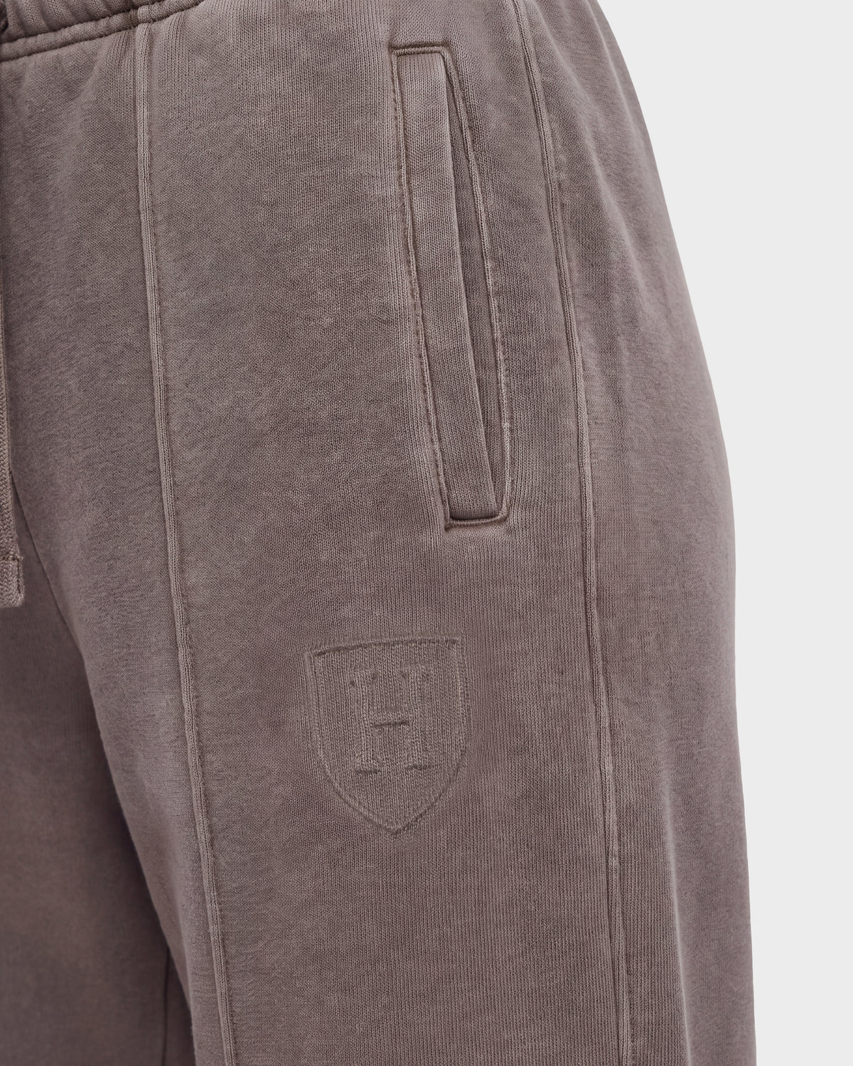 Close-up of the GAELLE HV - H526902 Taupe sweatpants by Rudsak, designed in a heavyweight jersey jogger style with a vertical side pocket and an embossed shield logo featuring the letter H on the upper thigh. The fabric appears soft and slightly textured, evoking the timeless appeal of classic Harvard apparel.