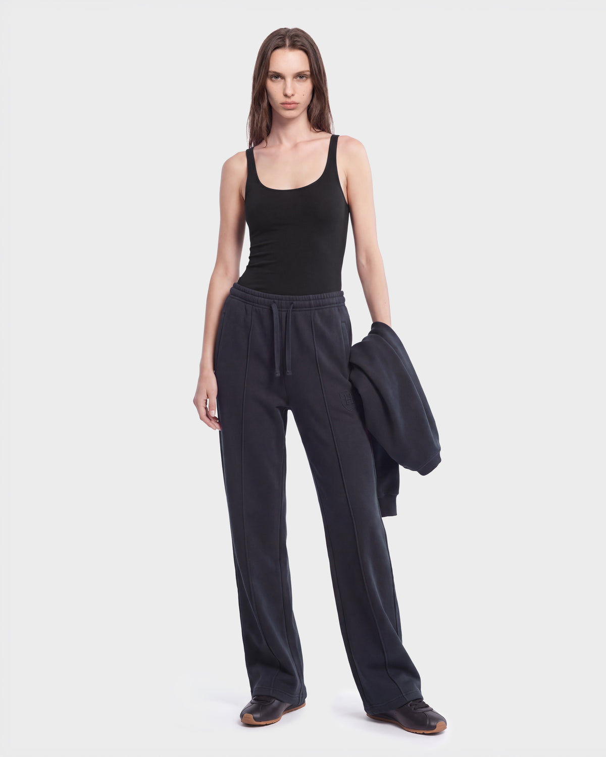 A person stands against a white background wearing a black tank top, Rudsak GAELLE HV - H526902 Titanium heavyweight jersey jogger pants, and black shoes, holding a matching black jacket in their left hand.