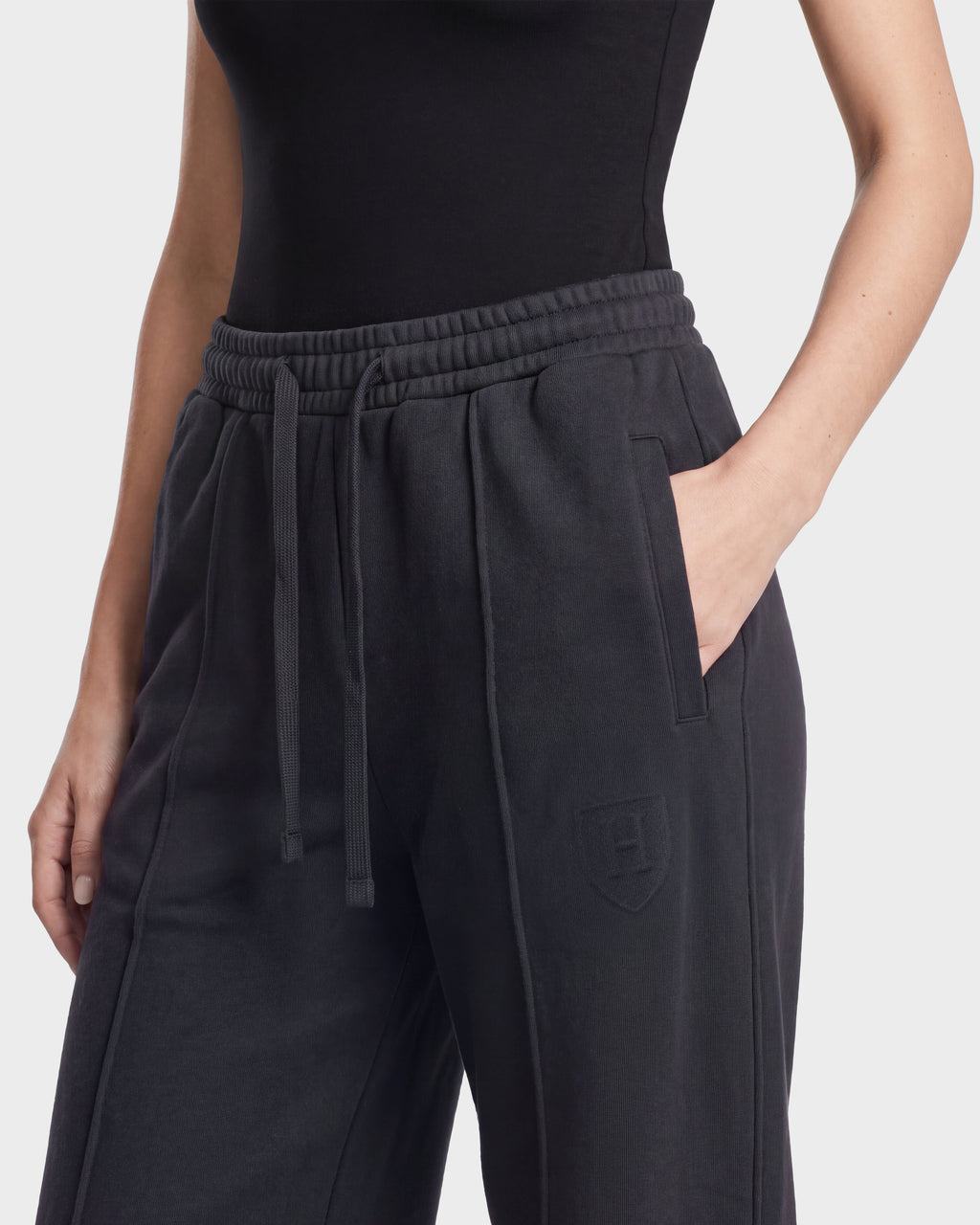 A person wears the Rudsak GAELLE HV - H526902 Titanium high-waisted, loose-fitting black sweatpants featuring a drawstring and shield logo, styled as heavyweight jersey joggers, paired with a fitted black top. One hand is in the pocket. The background is plain white.