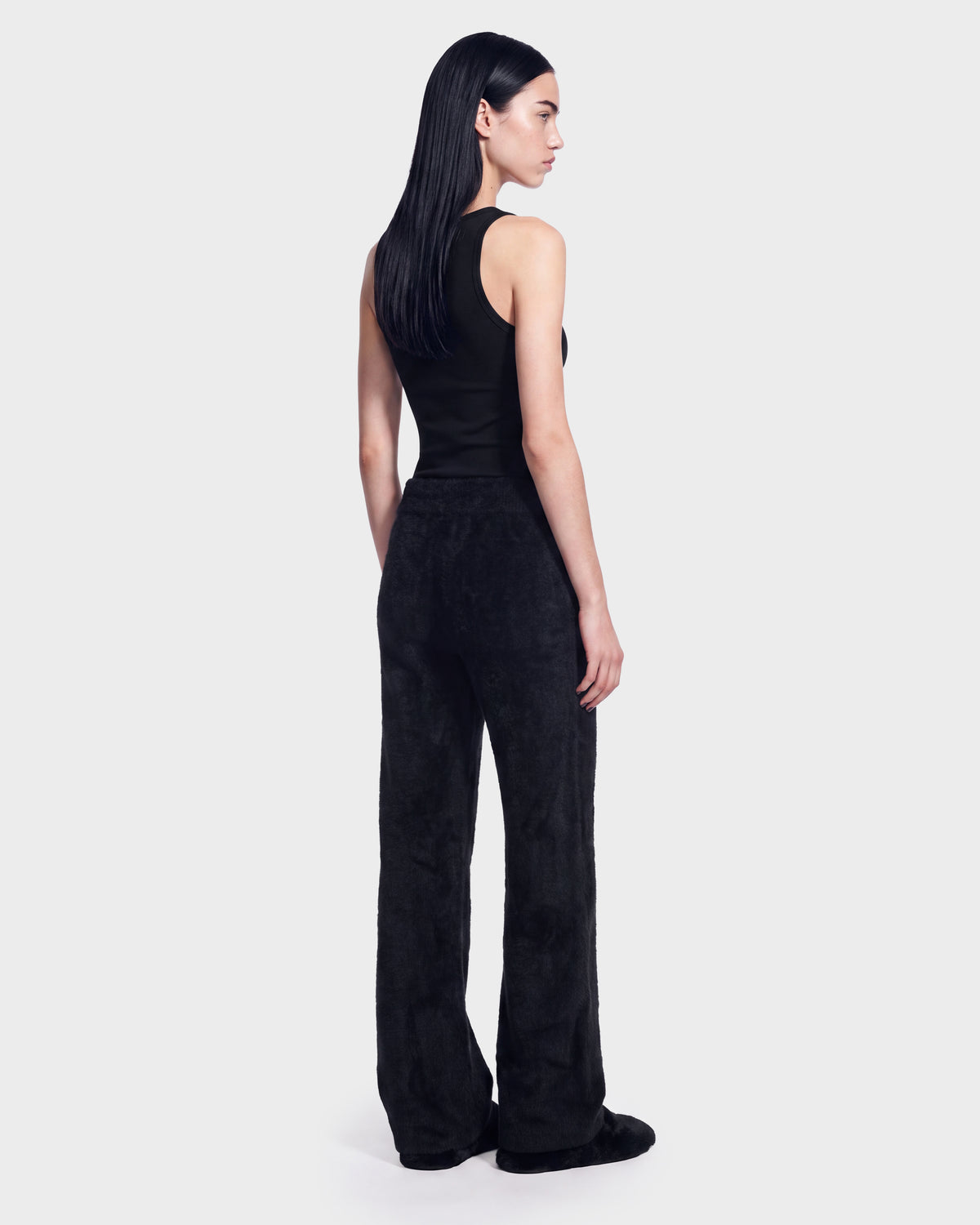 A woman with straight black hair is wearing the Rudsak GENESIS - 8526506 Black sleeveless top and matching black wide-leg pants, standing and facing slightly away against a plain light background.