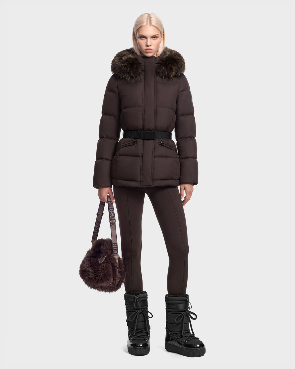 A model wearing a dark brown Rudsak GRACE FF - 8126535 Espresso goose down puffer coat with a faux-fur trimmed hood, a belt, and leggings, carrying a fluffy brown handbag and wearing black winter boots.
