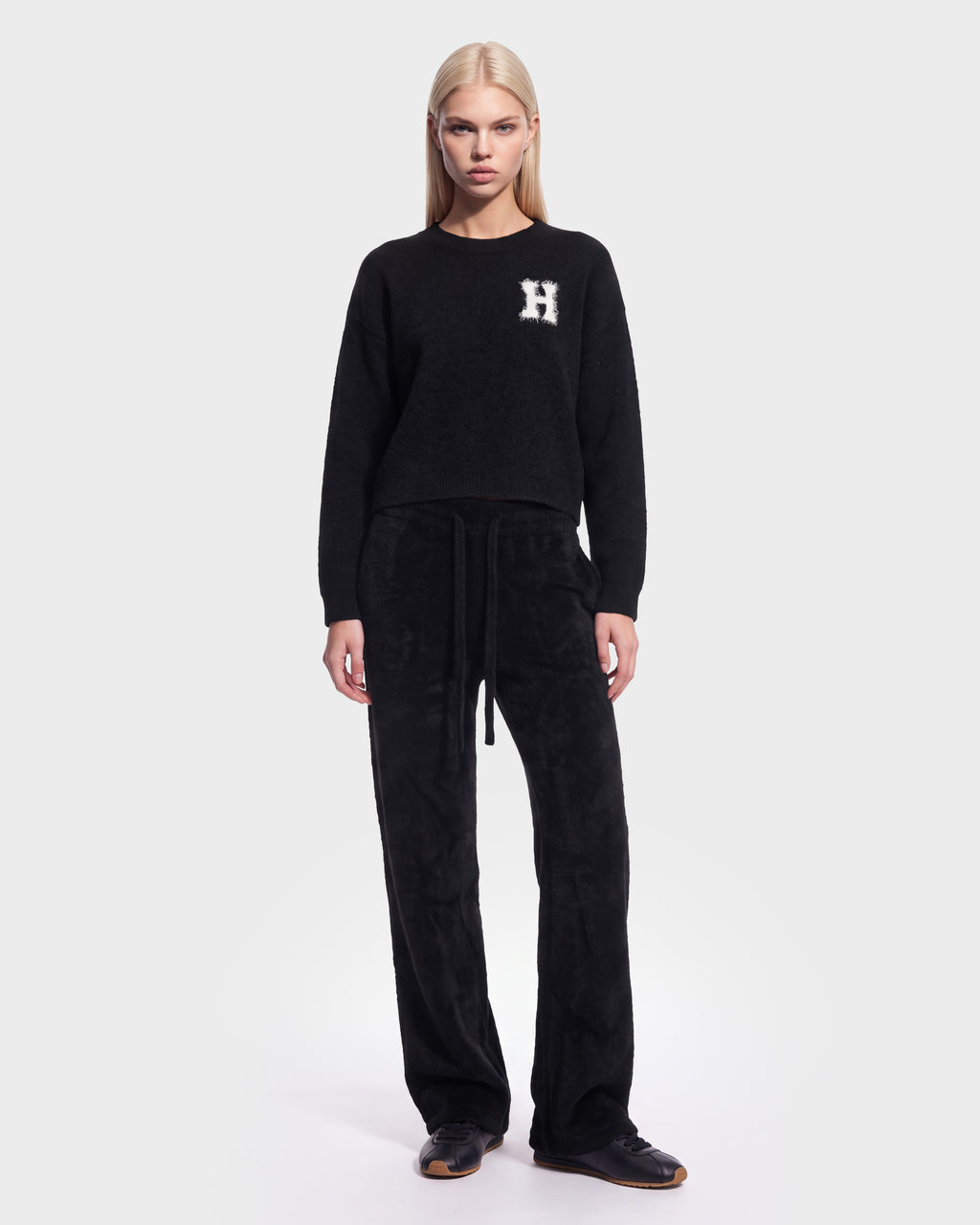 A woman with straight blonde hair stands facing forward against a plain white background, wearing the Rudsak HALLIE HV - H526506 Black crewneck sweater featuring a white “H” on the left chest and paired with black pants.