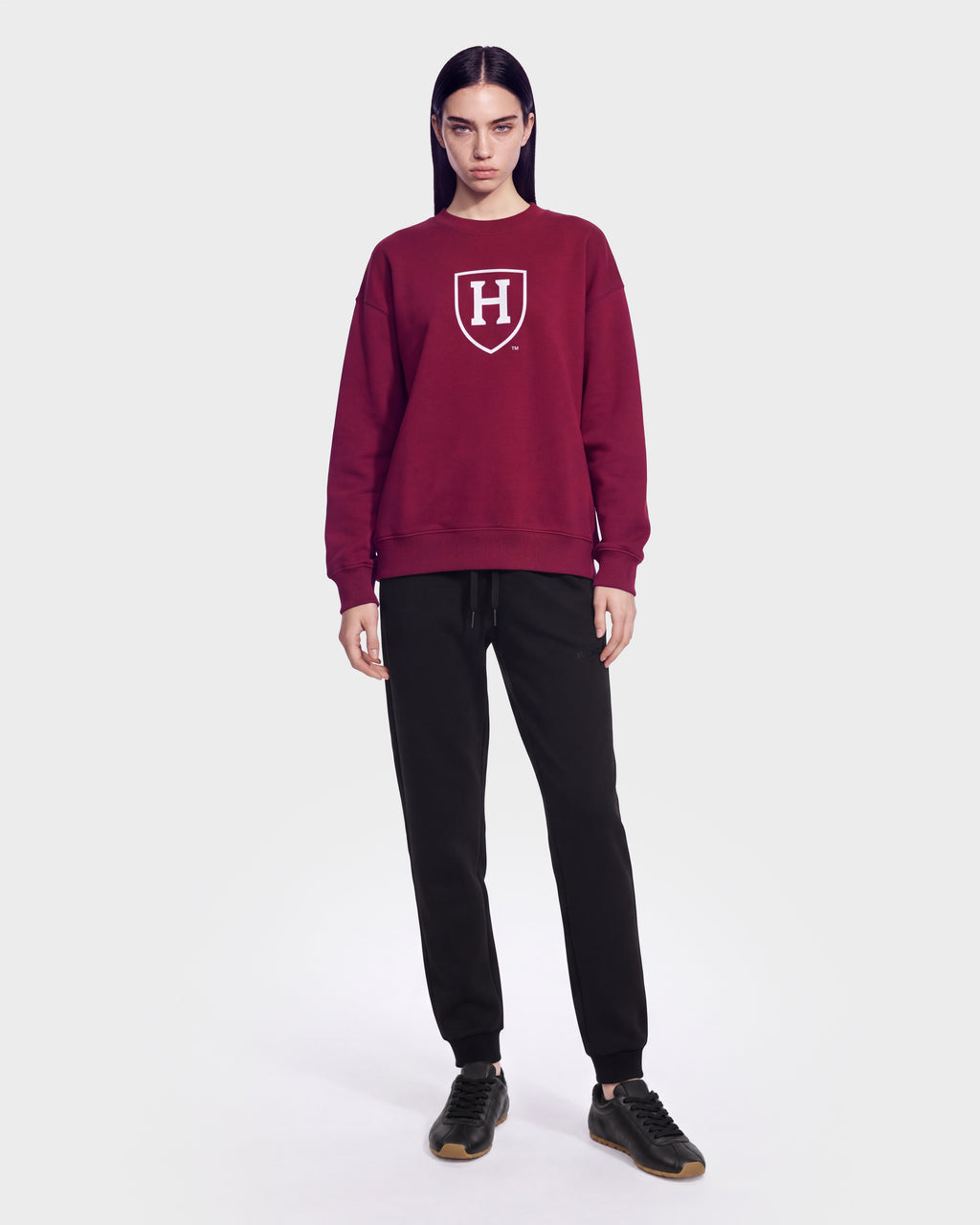 A person with long dark hair wears the Rudsak HARVARD ONE CRIMSON SWEATSHIRT 9524507, featuring a white H shield logo, paired with black pants and standing against a plain white background, embodying classic Ivy League style.