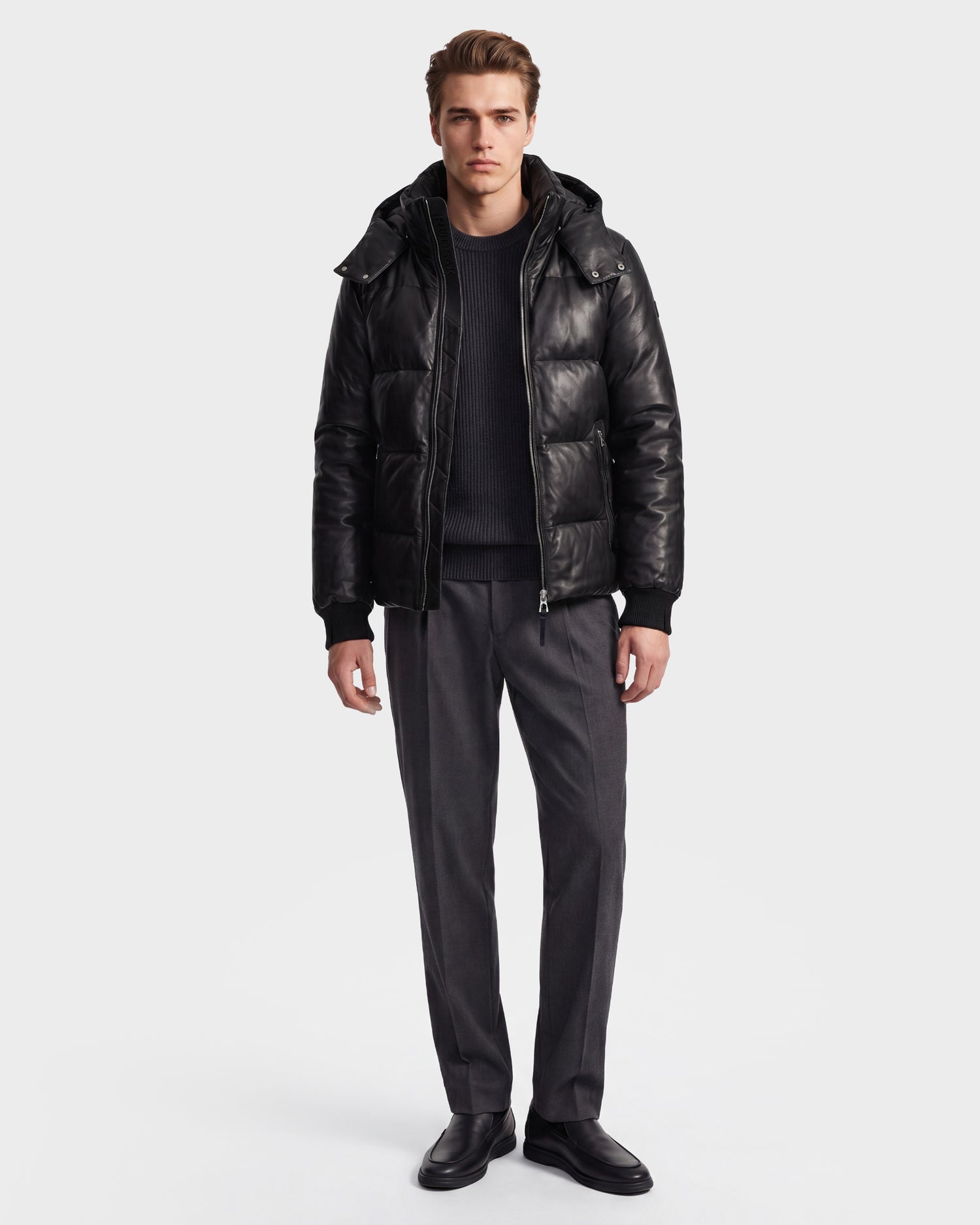 A man stands facing forward against a plain white background, wearing the Rudsak HARVEY X - 6125681 Black leather puffer jacket crafted with RDS-certified down, paired with a black sweater, gray trousers, and black shoes.