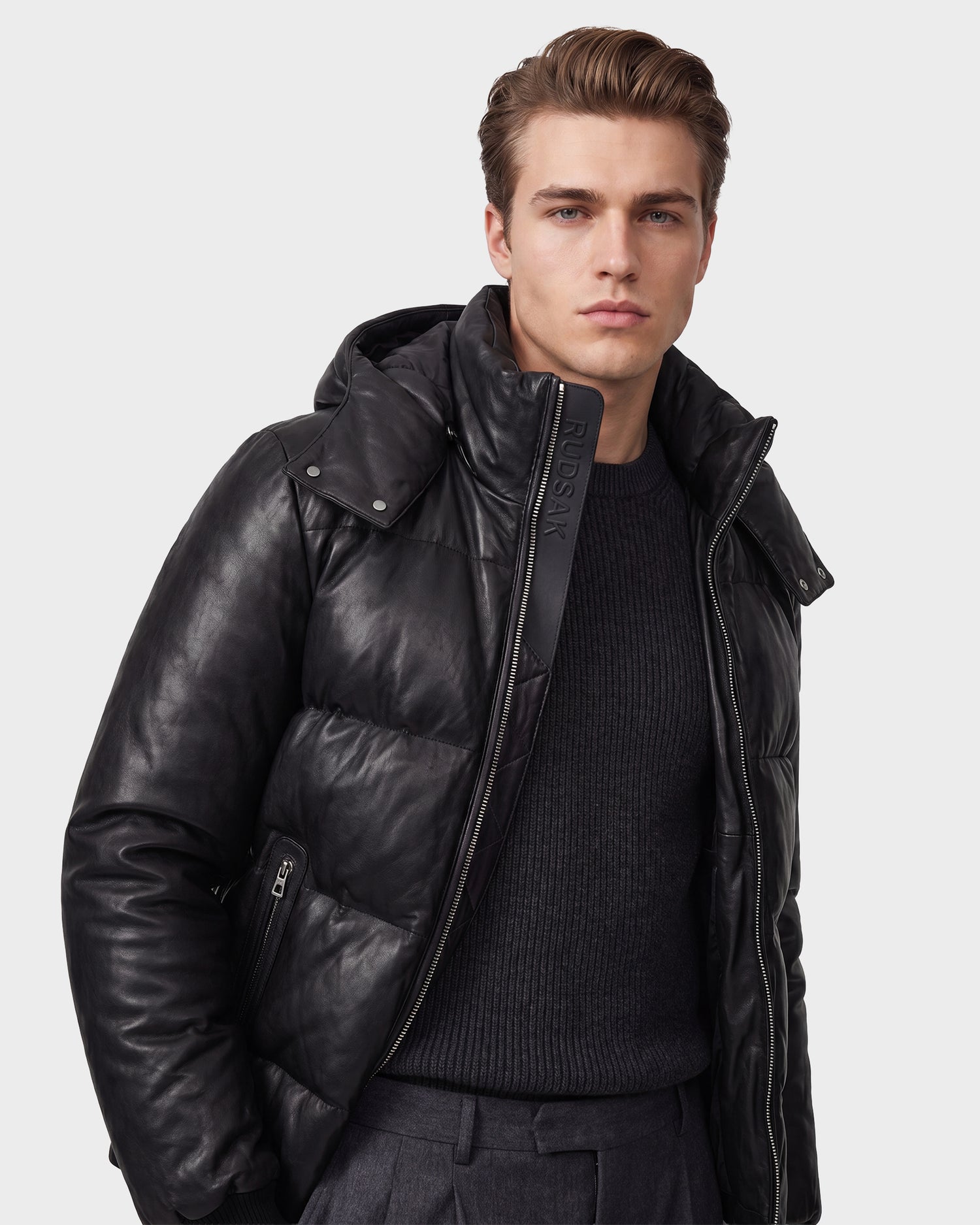 A man stands against a plain light background, looking straight ahead with a neutral expression. He is wearing the Rudsak HARVEY X - 6125681 black leather puffer jacket filled with RDS-certified down over a dark sweater and gray trousers.