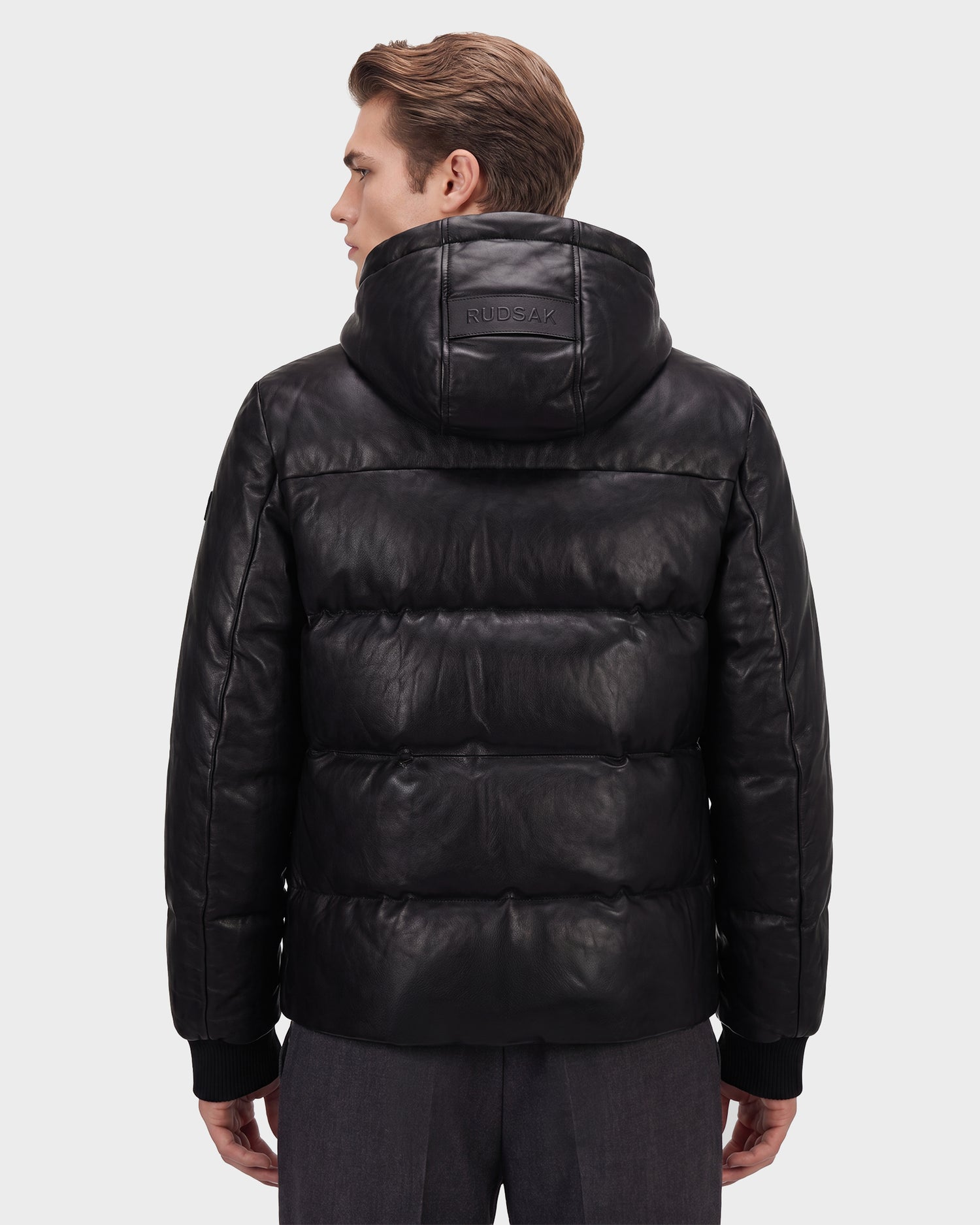 A man with light brown hair is seen from the back wearing the Rudsak HARVEY X - 6125681 Black leather puffer jacket with a hood, filled with RDS-certified down and featuring “RUDSAK” embossed on the hood. He stands against a plain light background.