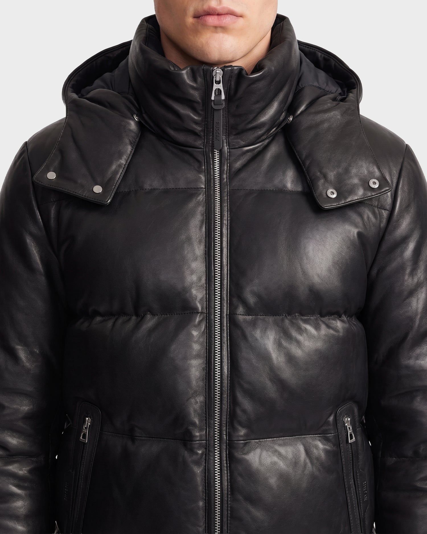 A person wears the HARVEY X - 6125681 Black puffer jacket by Rudsak, crafted from sleek black leather and insulated with RDS-certified down for extra warmth. The jacket features a zip-up front and snap buttons on the hood. The image is cropped at the shoulders and mid-face.