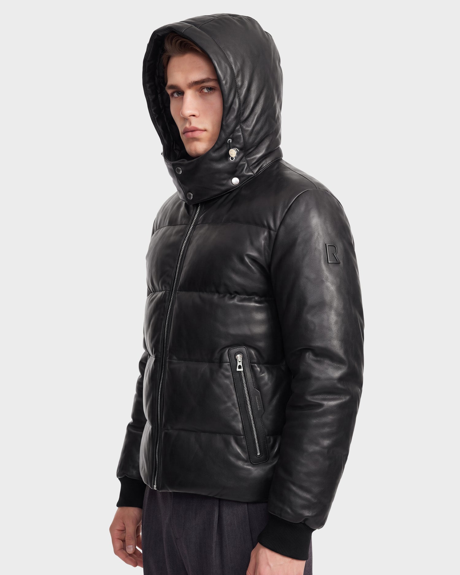 A young man in the Rudsak HARVEY X - 6125681 Black leather puffer jacket with a hood stands against a plain light gray background, looking serious. This men’s parka features zippered pockets, a high collar, and is filled with RDS-certified down for warmth and style.