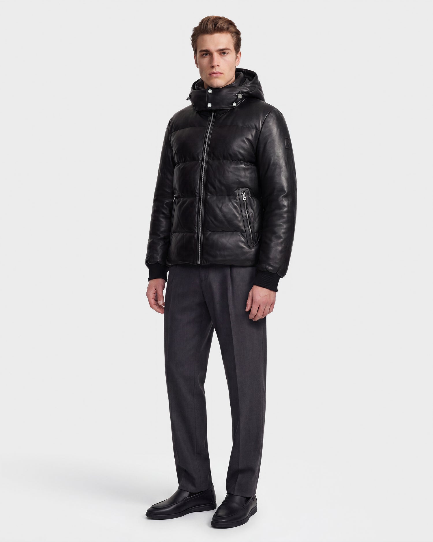 A man wearing the Rudsak HARVEY X - 6125681 Black leather puffer jacket with RDS-certified down, gray dress pants, and black shoes stands against a plain white background.
