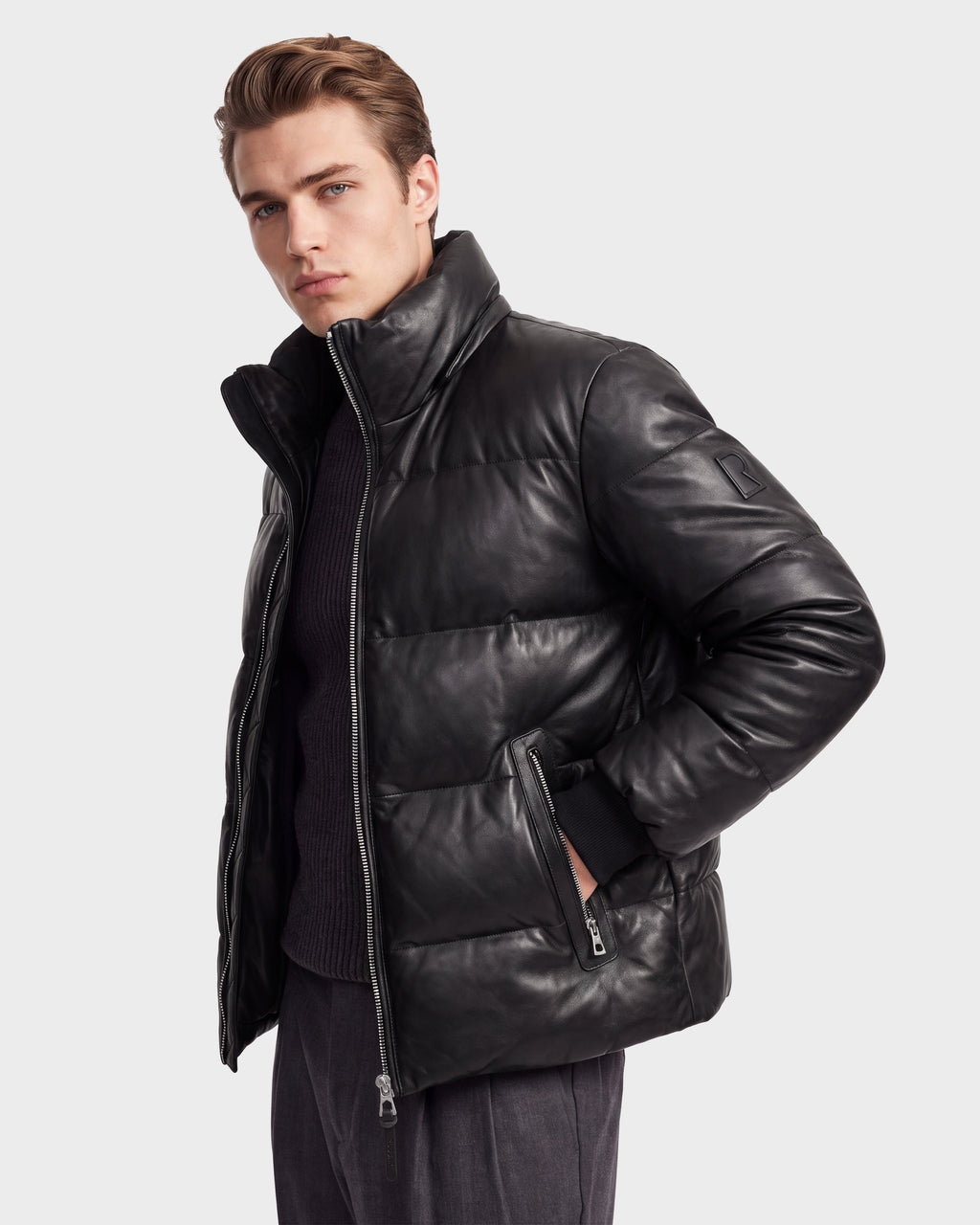 A man wearing the HARVEY X - 6125681 Black parka by Rudsak stands with one hand in his pocket, looking at the camera. This men’s leather parka features a high collar and zippered pockets, showcasing the signature style of Rudsak outerwear.