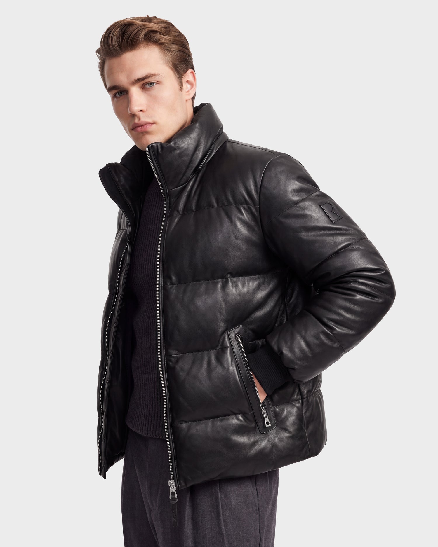 A man wears the Rudsak HARVEY X - 6125681 Black RDS-certified down leather puffer jacket, featuring a high collar and zippered pockets, as he stands against a plain white background and looks at the camera with a serious expression.