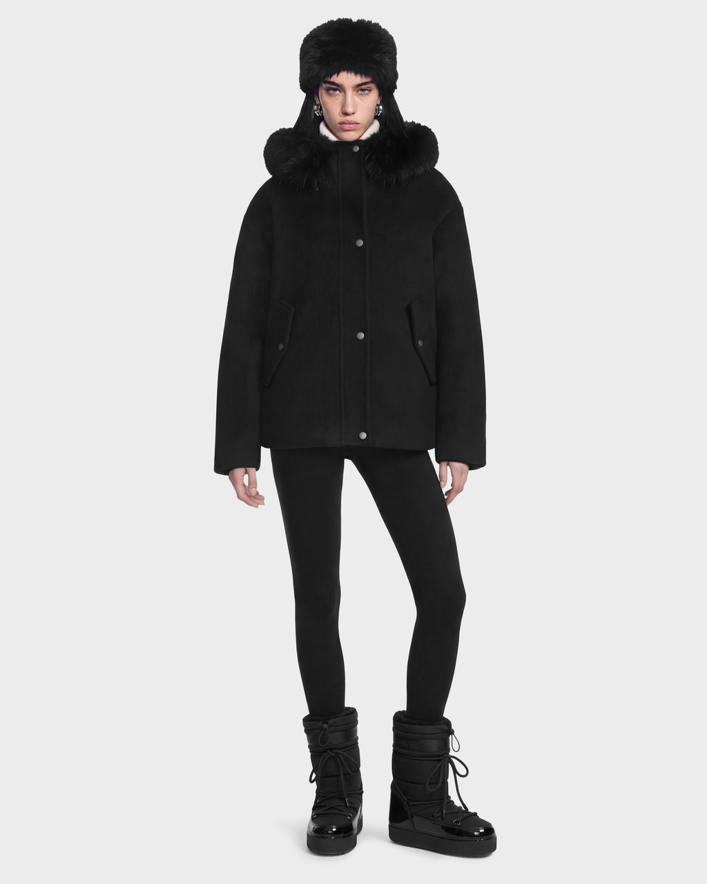 A person wears a Rudsak HAZEL - 8126981 Black wool-blend winter coat with a faux-fur trimmed hood, a matching fur hat, black leggings, and black winter boots.