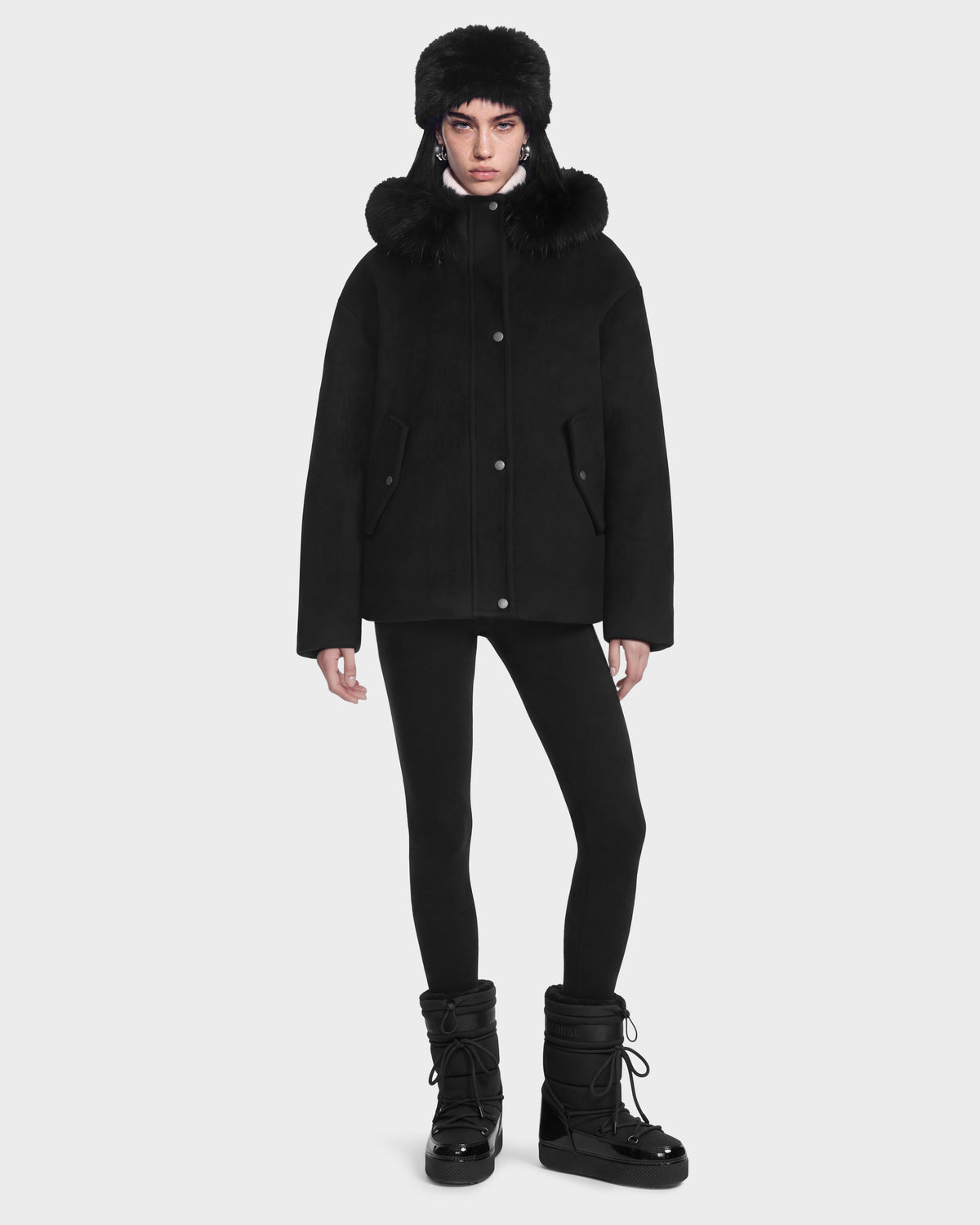 A person wears a Rudsak HAZEL - 8126981 Black wool-blend winter coat with a faux-fur trimmed hood, a matching fur hat, black leggings, and black winter boots.