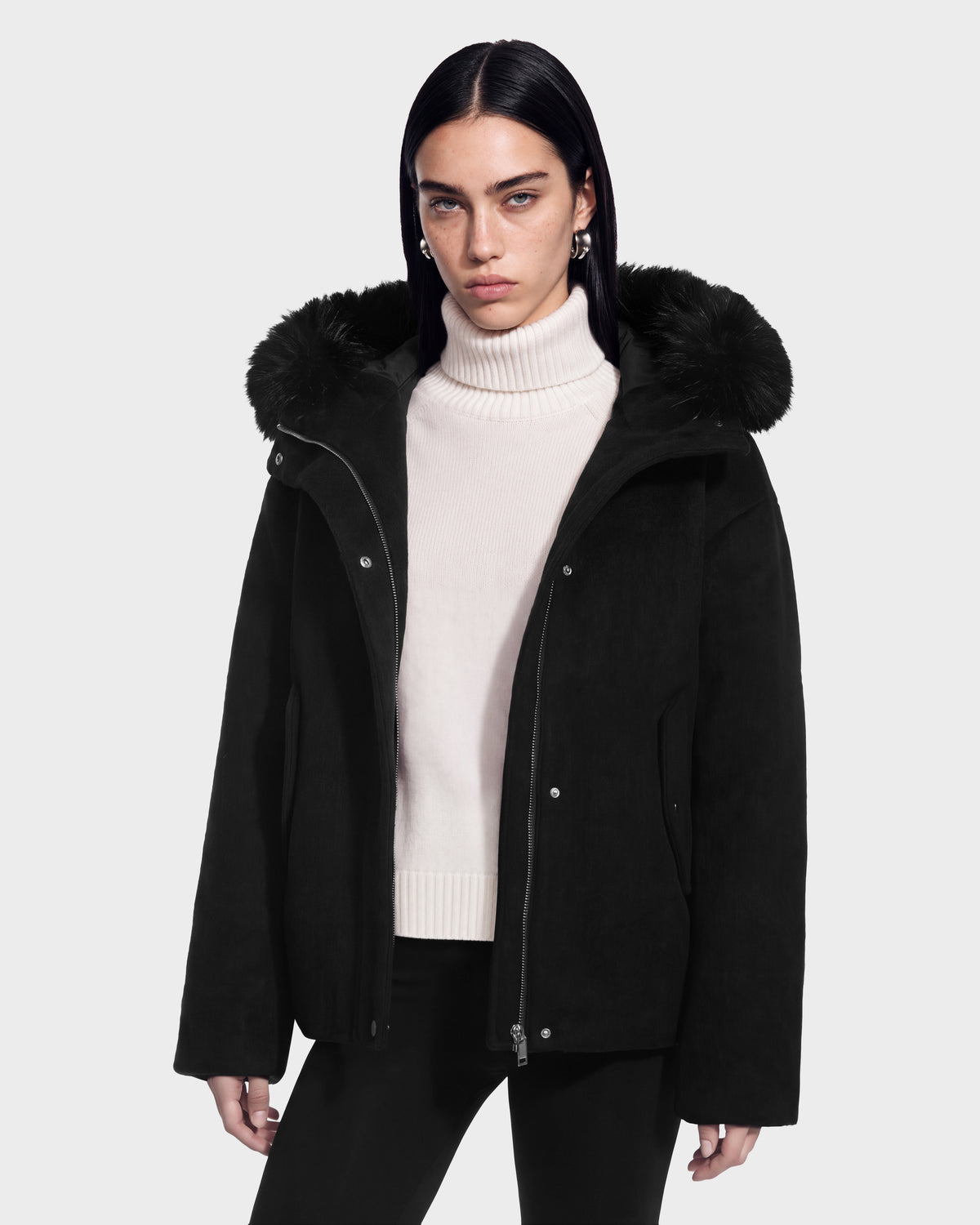 A person wearing a black Rudsak HAZEL 8126981 wool-blend winter coat with a faux-fur trimmed hood over a white turtleneck sweater.
