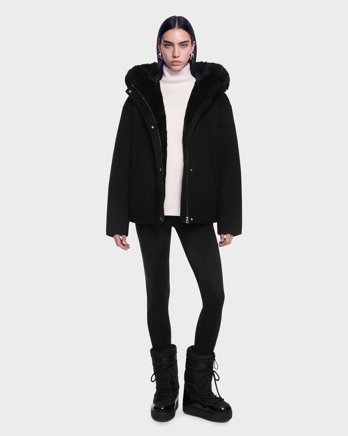 A model wearing a black HAZEL - 8126981 Black wool-blend winter coat with a faux-fur trimmed hood over a white turtleneck, black leggings, and black lace-up winter boots.