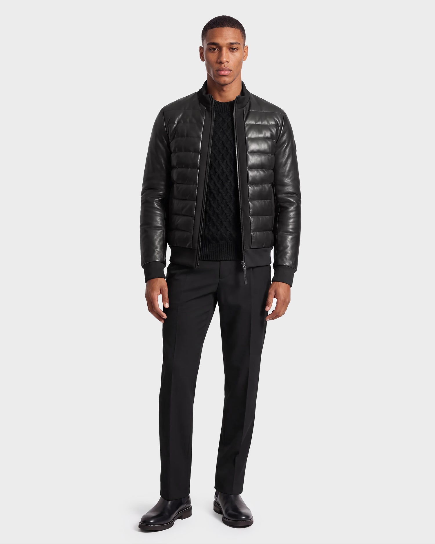 A man stands facing forward against a plain white background, wearing the Rudsak JIMMY X - 6124540 Black men's quilted leather down jacket, along with a black sweater, black pants, and black shoes.