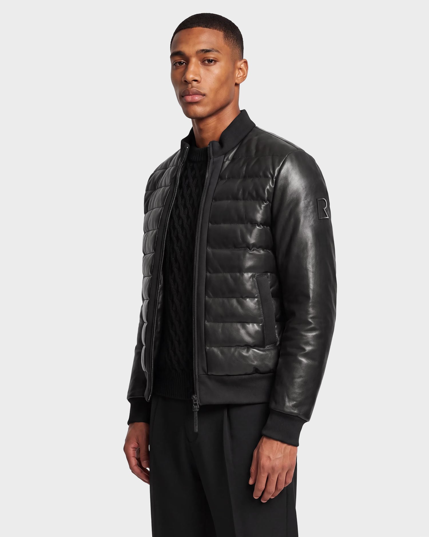 A man stands against a light background wearing the Rudsak JIMMY X - 6124540 Black quilted leather down jacket, paired with a black sweater and black pants, looking slightly to the side with a neutral expression.