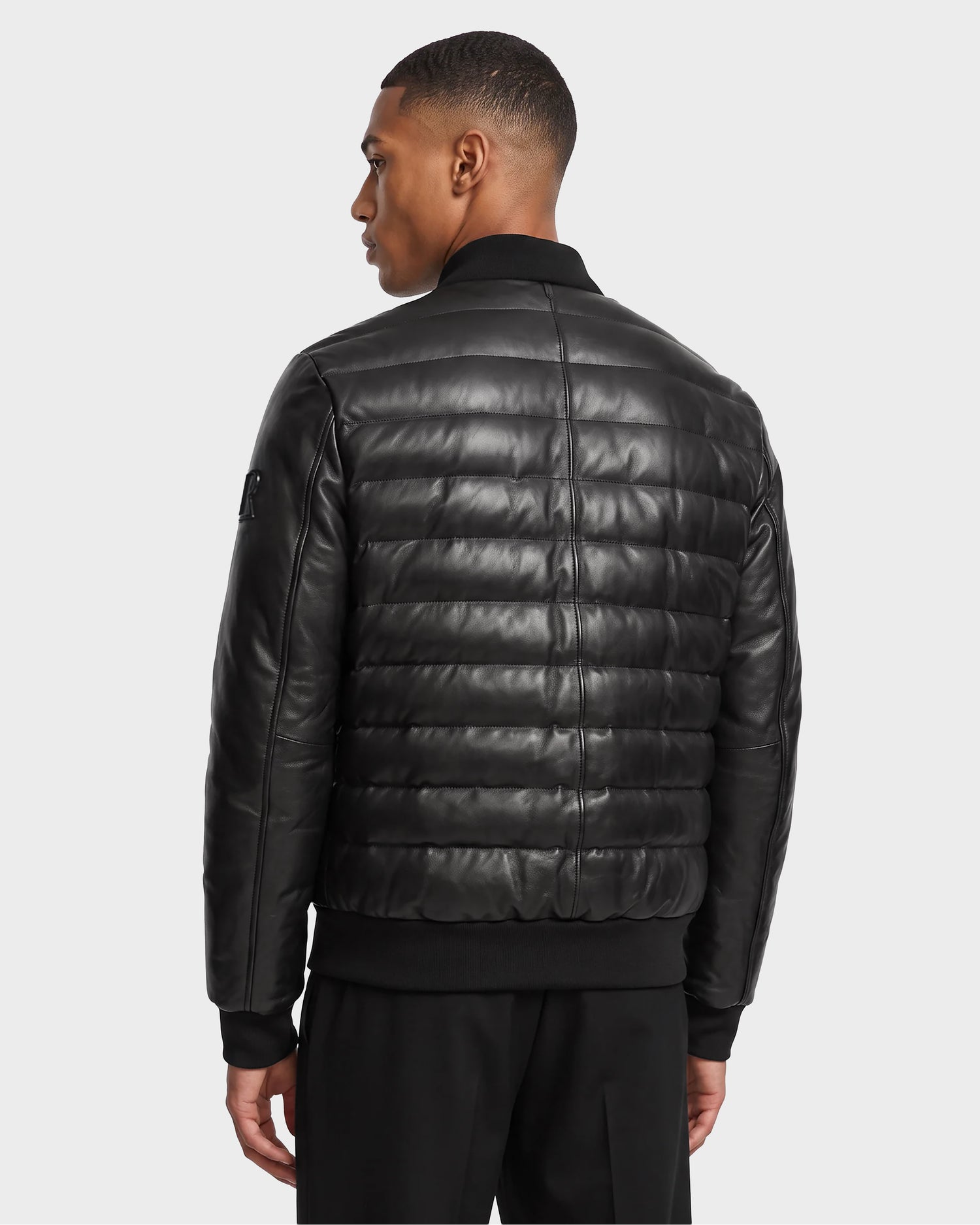 A man with short hair is seen from behind, wearing the Rudsak JIMMY X - 6124540 Black men’s quilted leather down jacket featuring ribbed cuffs and waistband, paired with black pants, standing against a plain light gray background.