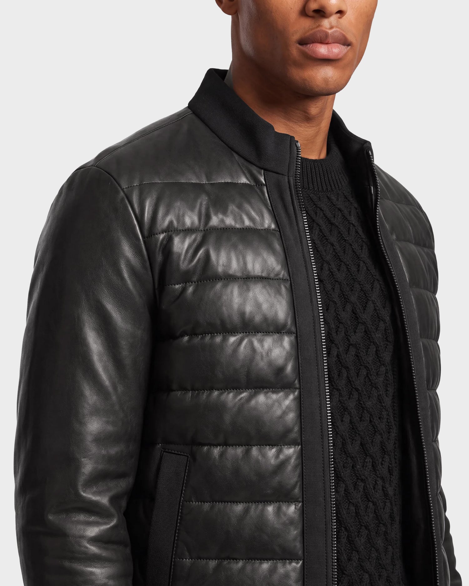 A man wearing the Rudsak JIMMY X - 6124540 Black men's quilted leather down jacket over a textured black sweater, standing slightly turned to the side against a plain light background. Only the lower half of his face is visible.