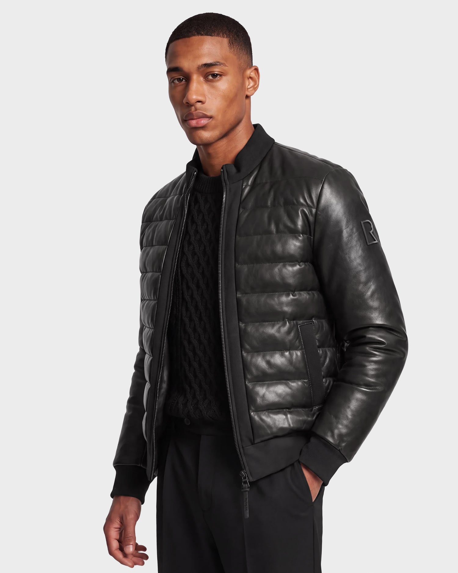 Wearing the Rudsak JIMMY X - 6124540 Black, a luxurious men's quilted leather down jacket crafted with RDS-certified down, a man stands against a plain background over a black cable-knit sweater and black pants, looking at the camera.