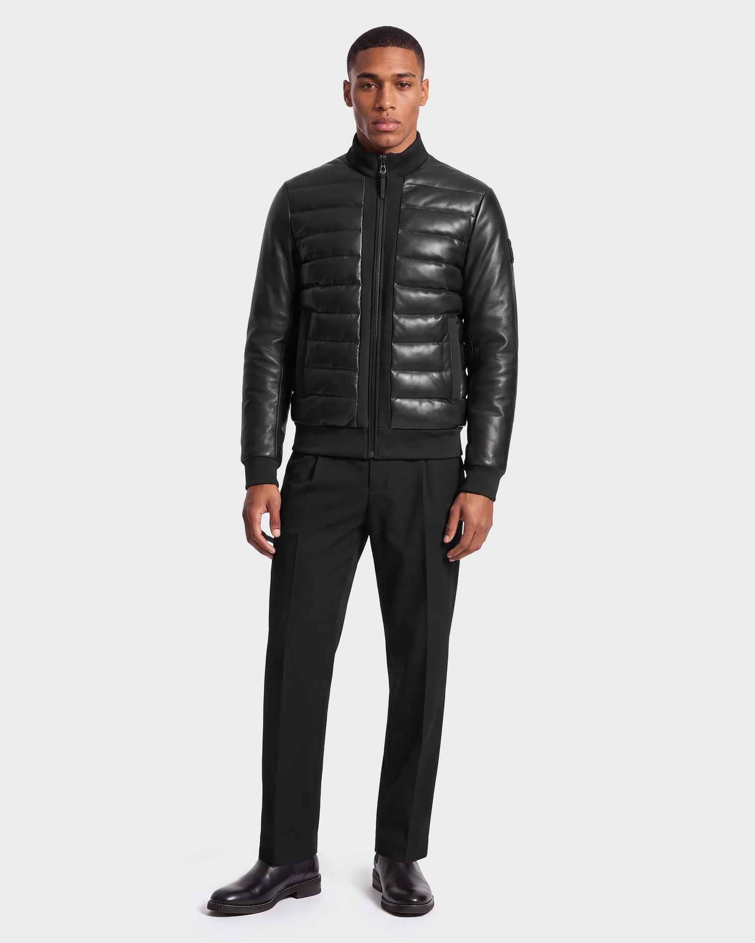 A man stands facing forward wearing the Rudsak JIMMY X - 6124540 Black men’s quilted leather down jacket, paired with black pants and black boots against a plain light background.