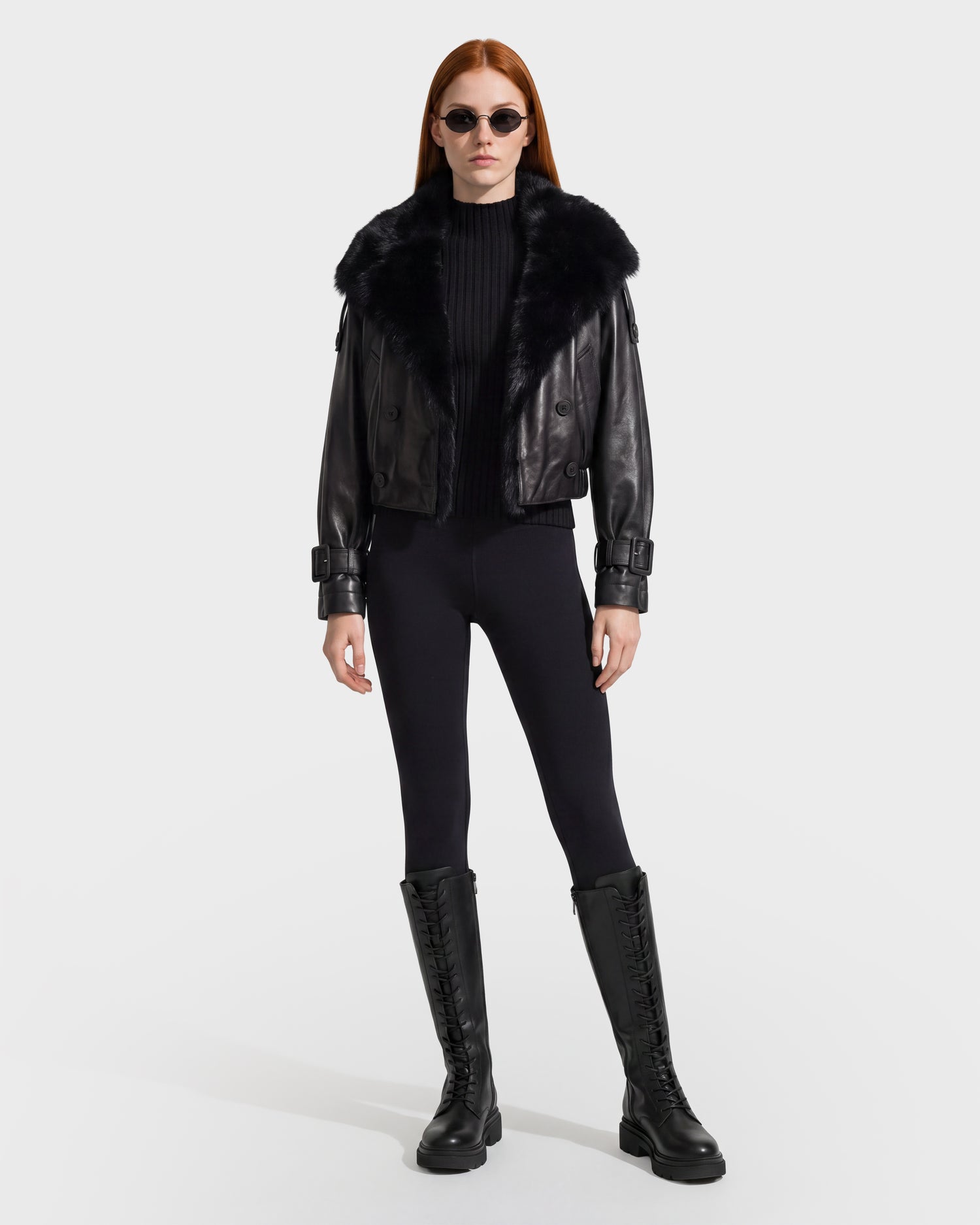 A woman with long red hair is wearing the Rudsak JANELLE - 8126936 Black double-breasted faux fur collar jacket, styled with a black ribbed top, black leggings, lace-up knee-high boots, and round sunglasses as she stands against a plain white background.