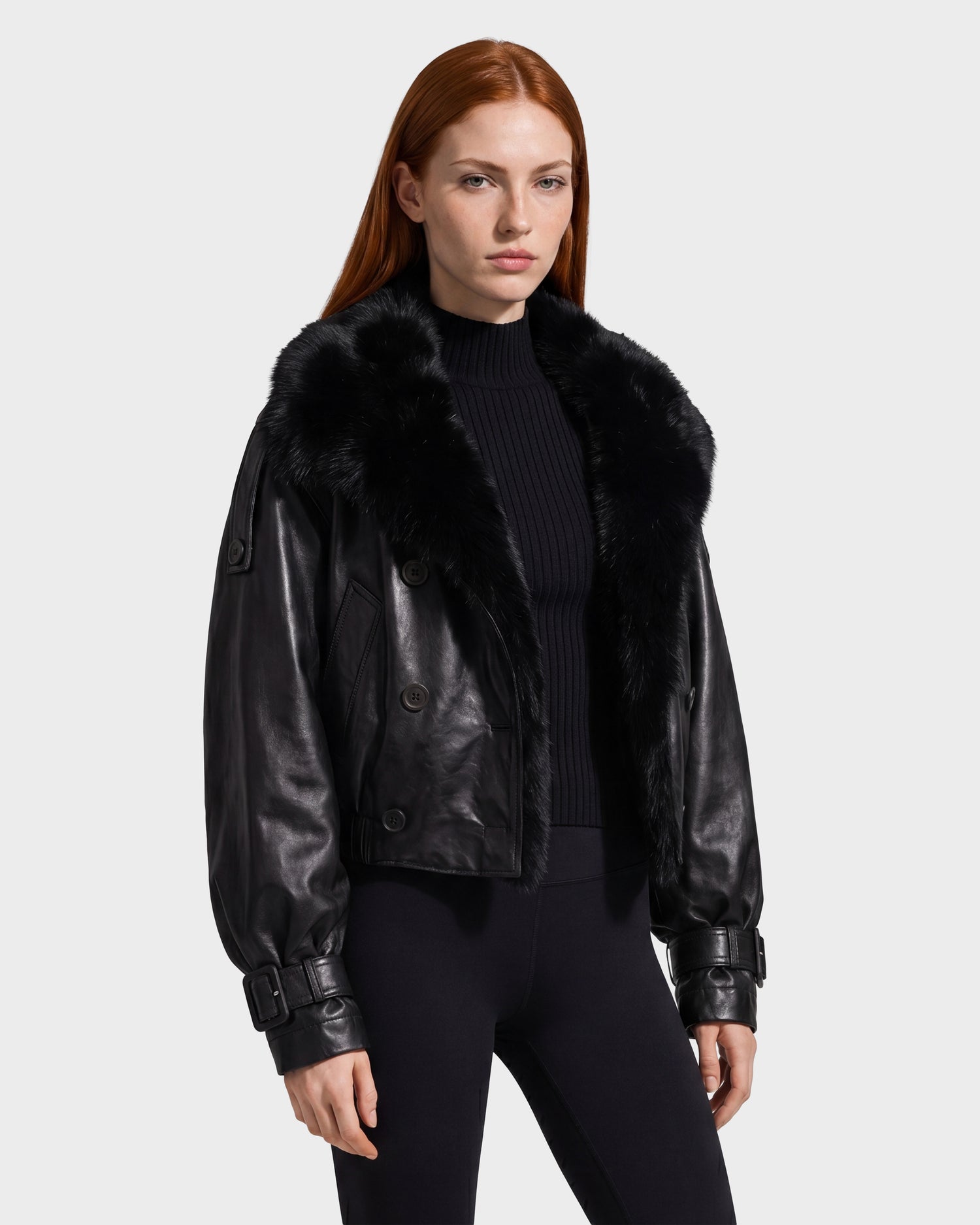 A woman with long red hair models the Rudsak JANELLE - 8126936 Black double-breasted leather bomber jacket with a faux fur collar, layered over a ribbed black top and black pants, standing against a plain light background.