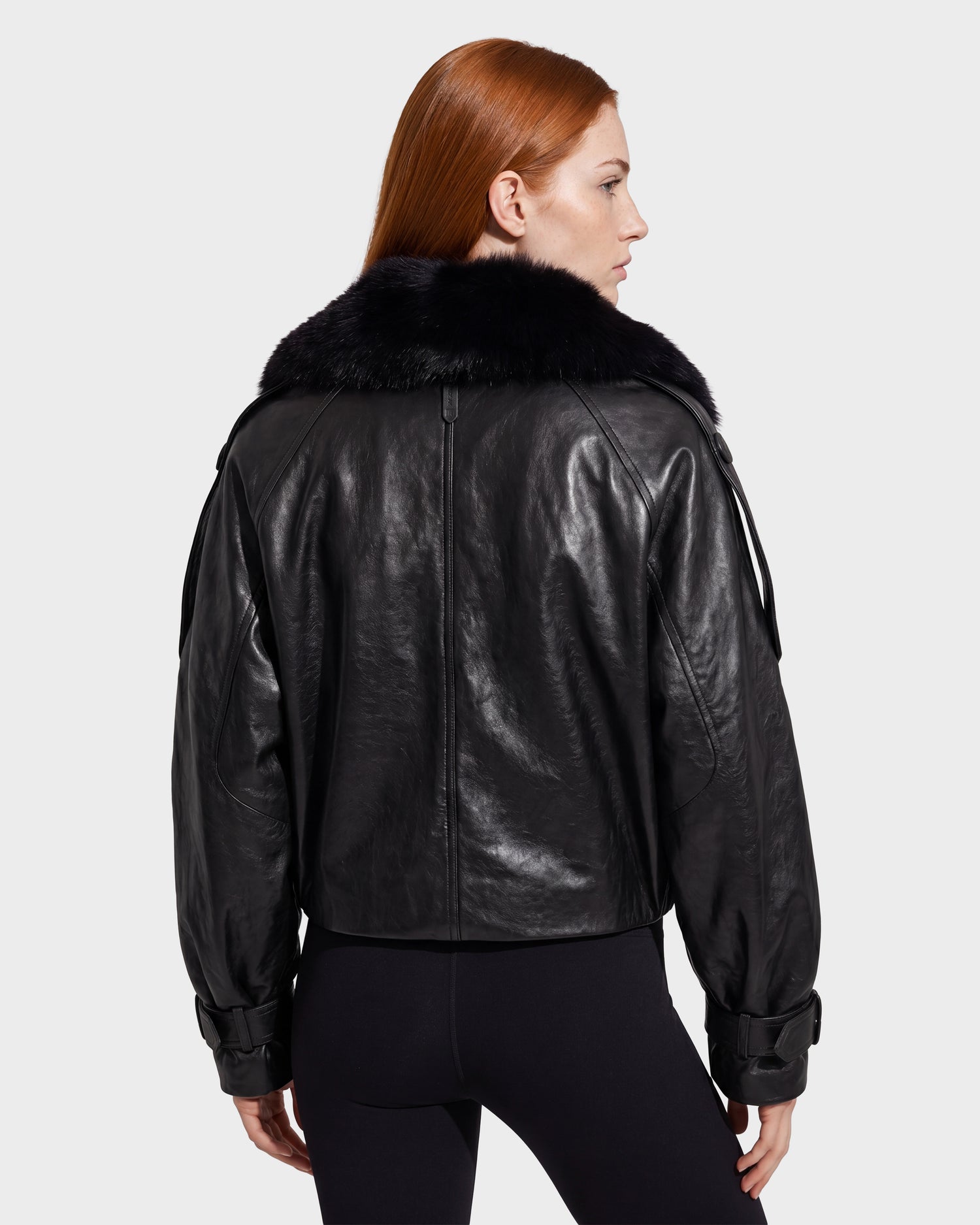 A woman with long red hair models the Rudsak JANELLE - 8126936 Black double-breasted leather jacket, which features a large faux fur collar and is shown from the back. She pairs it with black leggings against a plain light background.