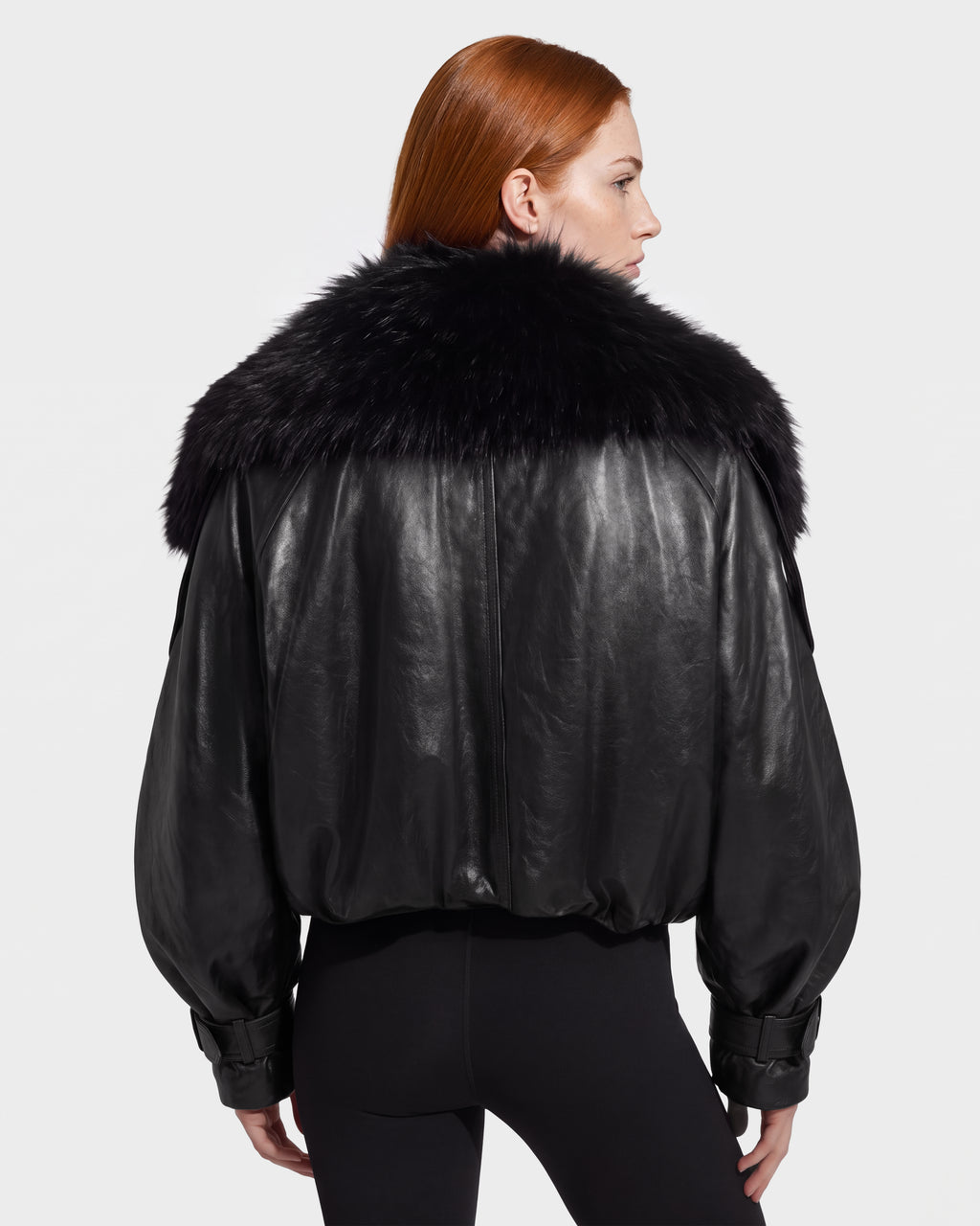 A person with long red hair wears the Rudsak JANELLE - 8126936 Black leather bomber jacket, distinguished by its large, fluffy faux fur collar. Shown from the back against a plain light background, this look embodies effortless city dressing.