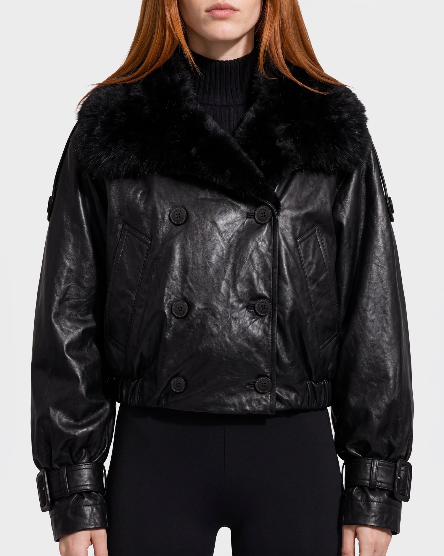 A person wearing the Rudsak JANELLE - 8126936 Black leather bomber jacket, featuring a large faux fur collar, button details, and belt accents on the cuffs, paired with black pants.
