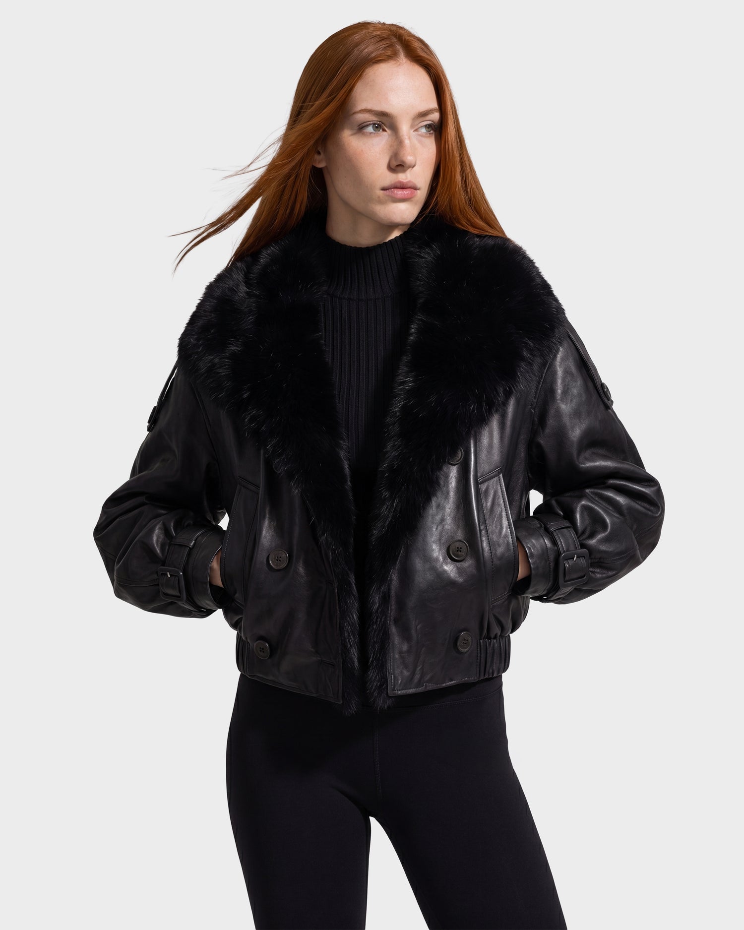 A woman with long red hair is wearing the Rudsak JANELLE - 8126936 Black leather bomber jacket, which features a large faux fur collar, layered over a black top and black pants. She stands with her hands in her pockets against a plain light background.