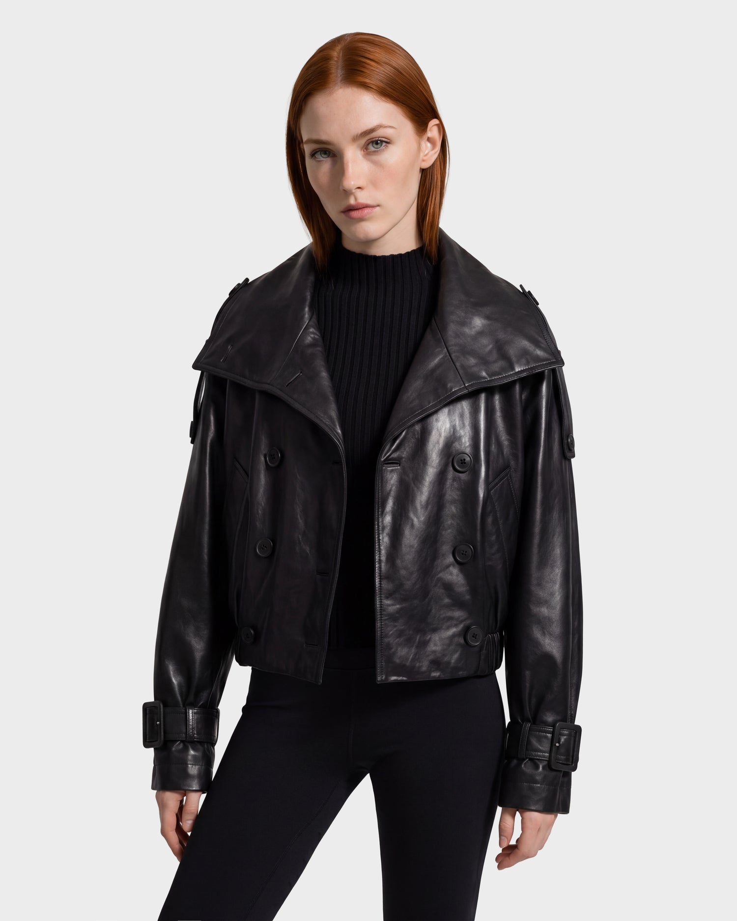 A woman with straight red hair wears the JANELLE - 8126936 Black double-breasted leather jacket by Rudsak, featuring large lapels, buckle details, and a sleek faux fur collar. She pairs it with a black ribbed sweater and black pants while standing against a plain light background.