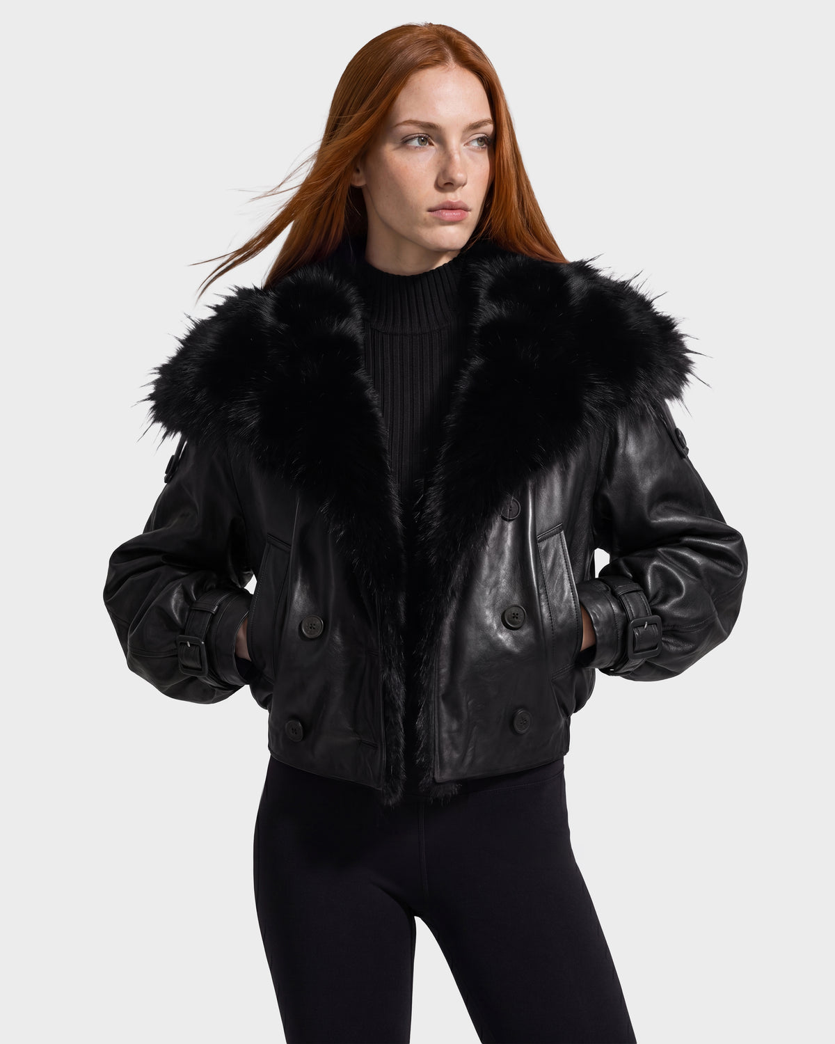 A woman with long red hair wears the Rudsak JANELLE - 8126936 Black leather bomber jacket, distinguished by its large faux fur collar, styled with black leggings and a ribbed black sweater for a chic city look as she stands against a plain light background.