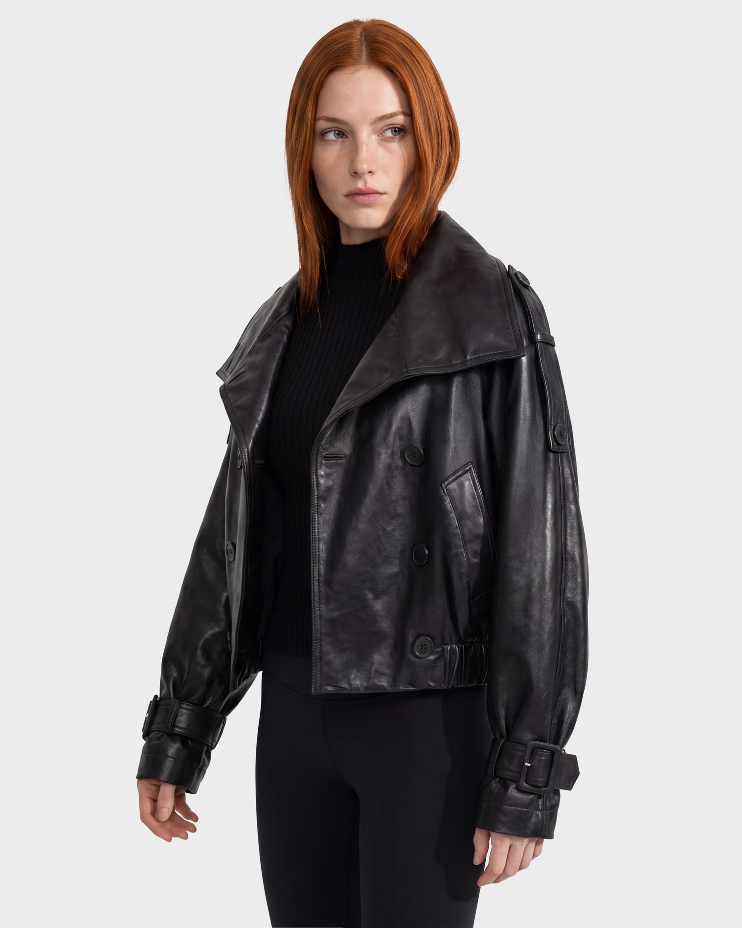 A woman with straight red hair wears the Rudsak JANELLE - 8126936 Black leather bomber jacket with a faux fur collar over a black top and leggings, standing against a plain light gray background and looking slightly to her left.
