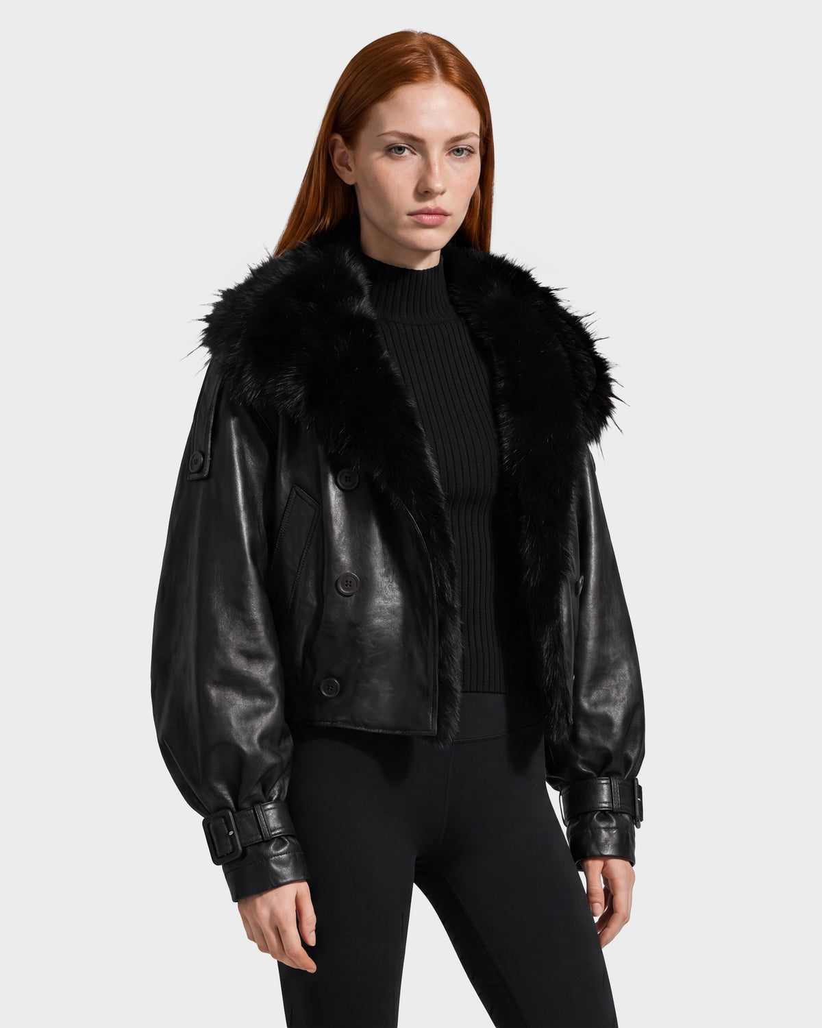 A woman with long red hair wears the Rudsak JANELLE - 8126936 Black leather bomber with a luxe faux fur collar, styled over a black ribbed turtleneck and black pants, exemplifying city dressing as she stands against a plain light gray background.