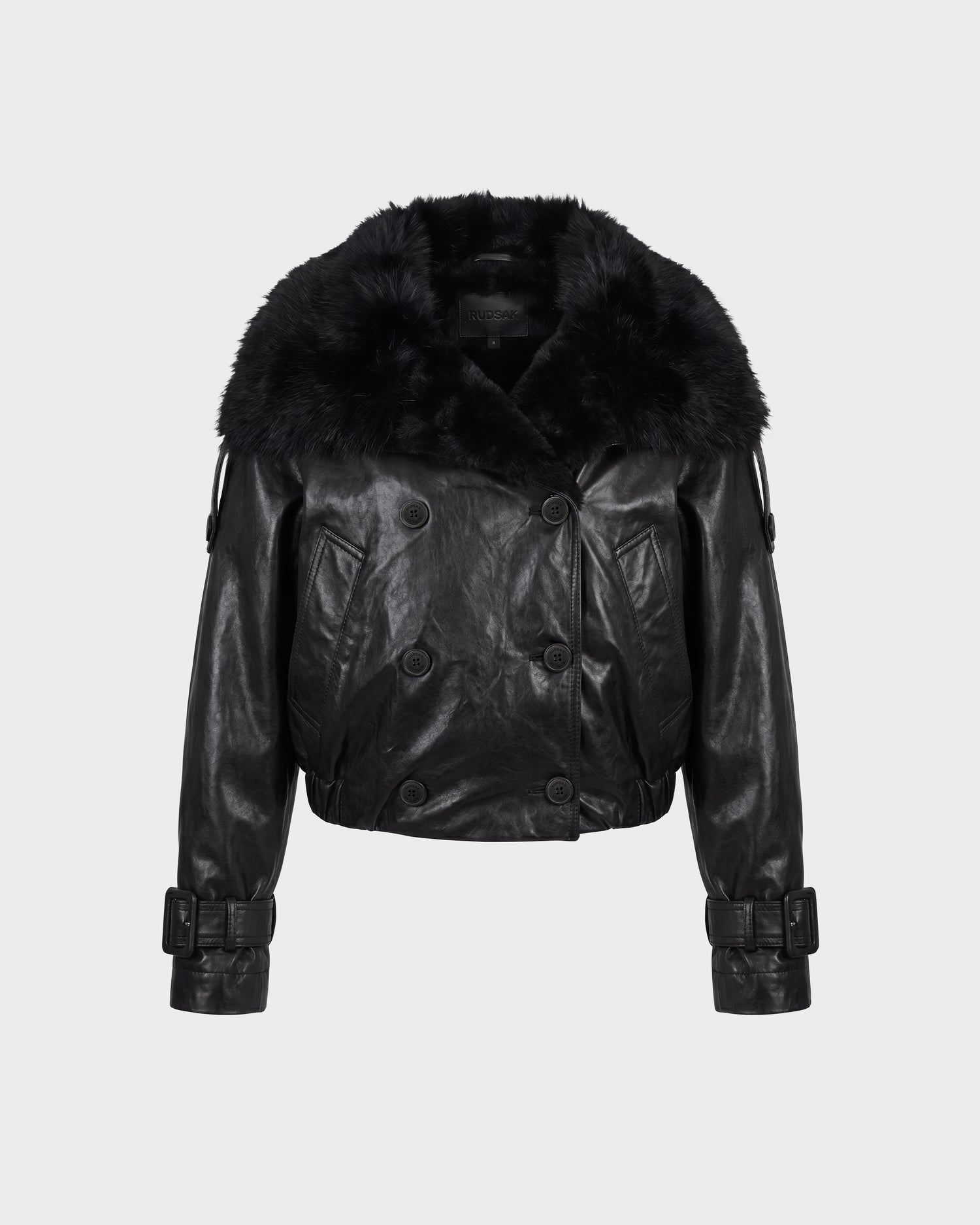 The JANELLE - 8126936 Black bomber jacket by Rudsak features black leather, a wide fluffy faux fur collar, double-breasted buttons, belted cuffs, and a cropped fit, displayed against a plain light background.