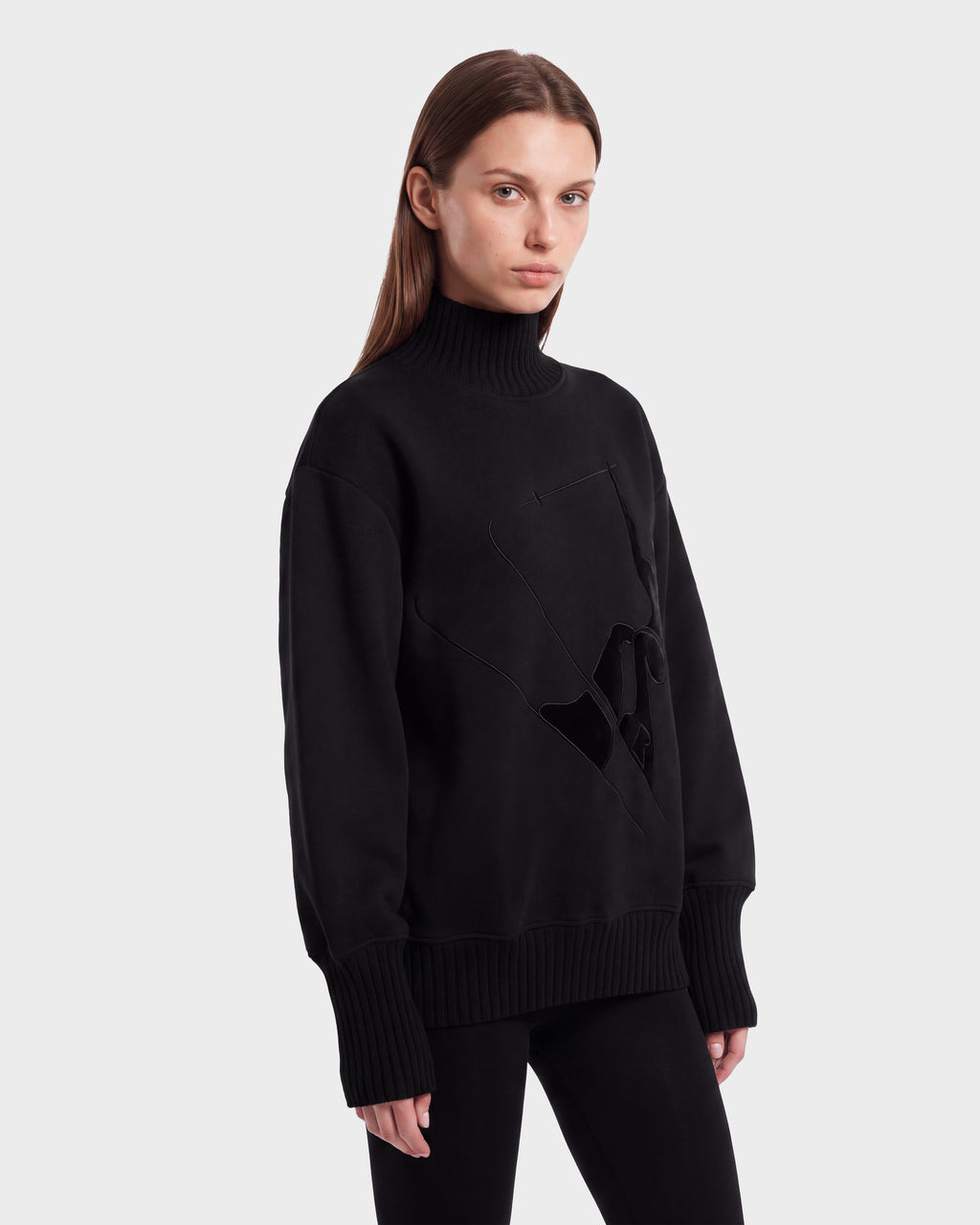 A woman with straight brown hair models the Rudsak JOYCE E - 8526538 Black turtleneck sweater, featuring textured detailing, paired with black pants and posed against a plain white background.