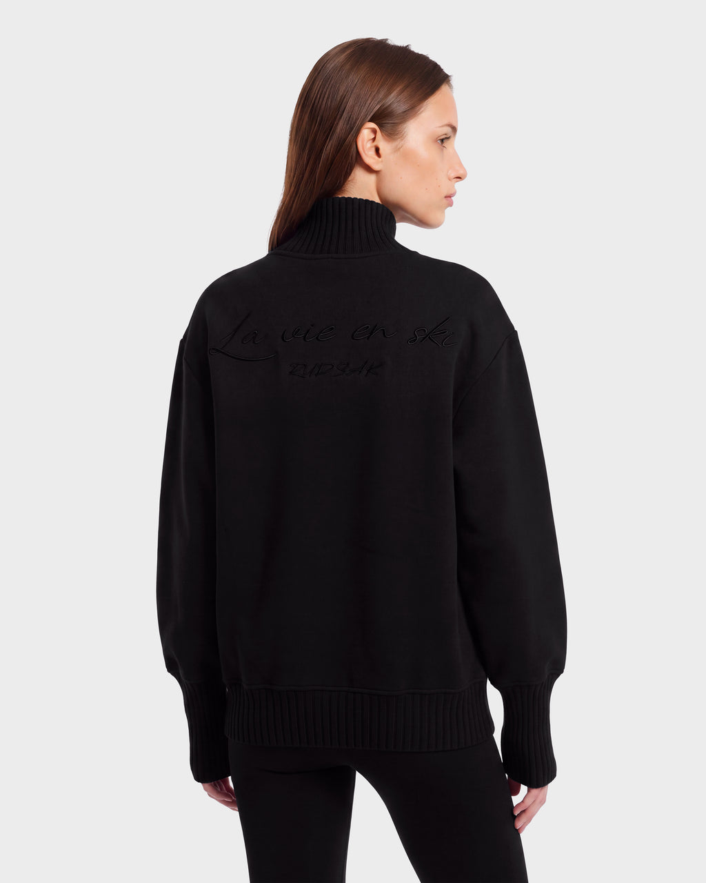A woman with straight brown hair wears the Rudsak JOYCE E - 8526538 Black turtleneck sweater, featuring embossed text on the back, paired with black pants. She stands against a plain light background, looking to her left.
