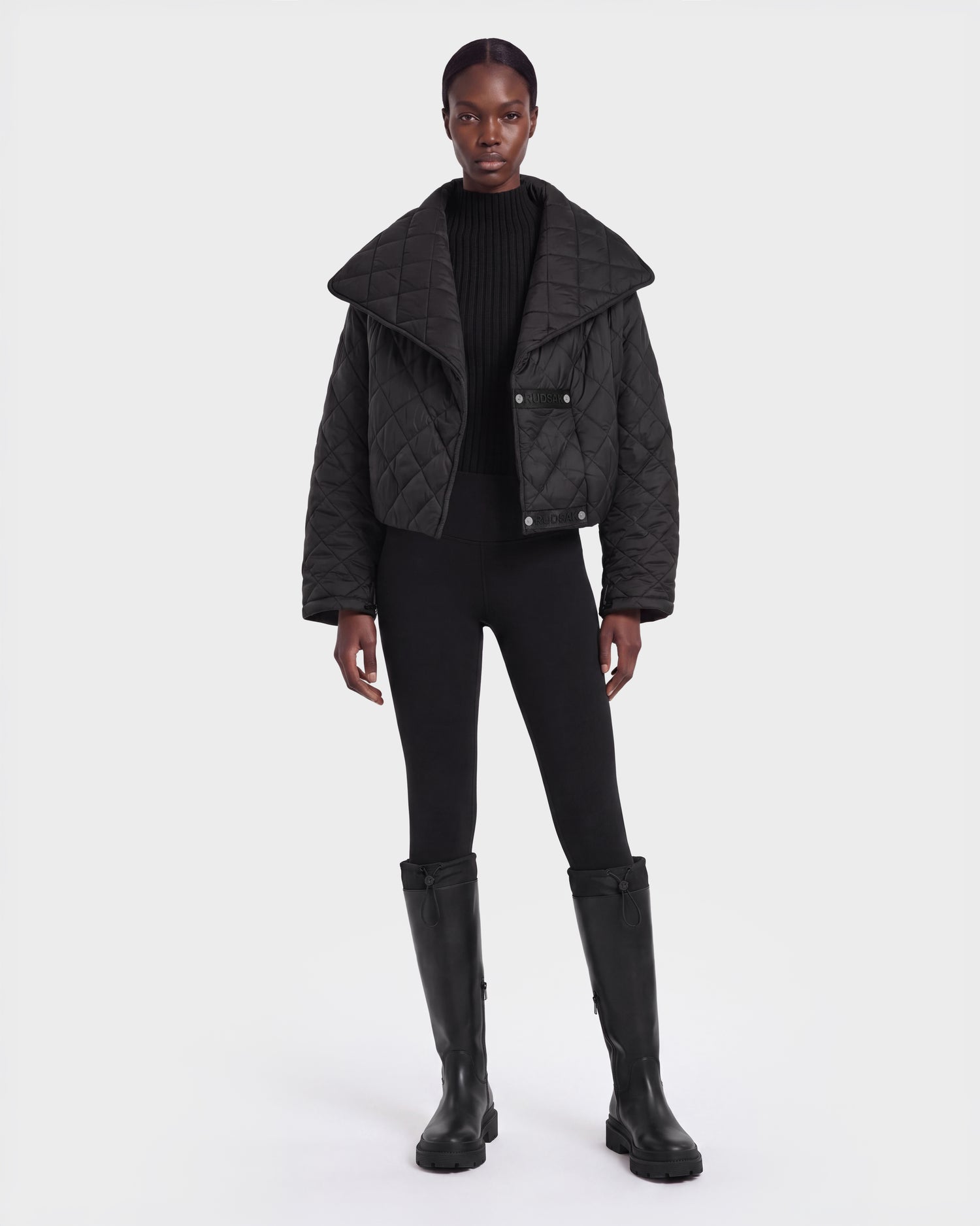 A person stands facing forward against a plain white background, wearing the Rudsak KALEE - 8124005 Black women's quilted light jacket with a large collar, paired with a black ribbed sweater, black leggings, and knee-high black boots.