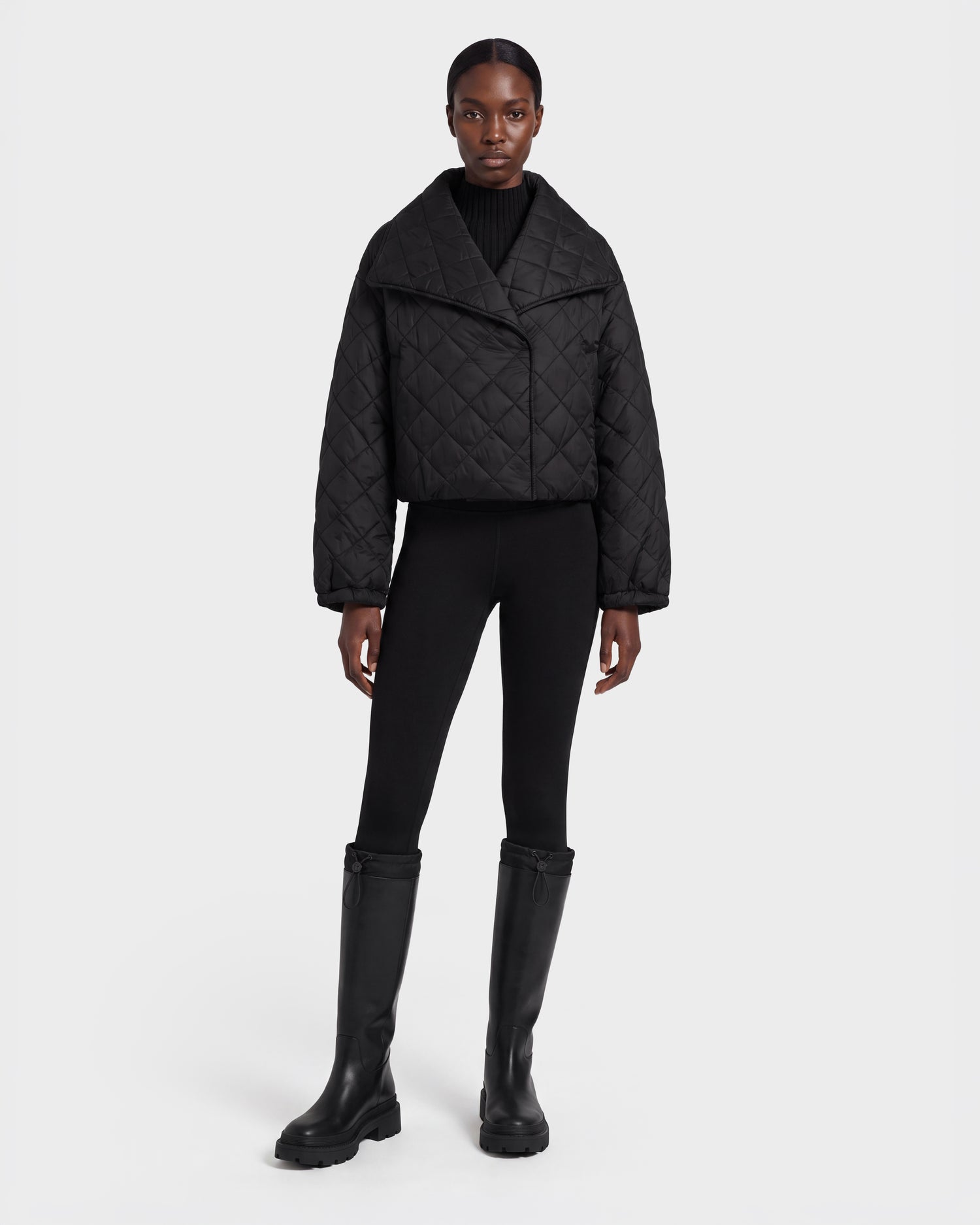 A person stands facing forward, wearing the Rudsak KALEE - 8124005 Black women's quilted light jacket paired with a black turtleneck, black leggings, and knee-high black boots against a plain white background.
