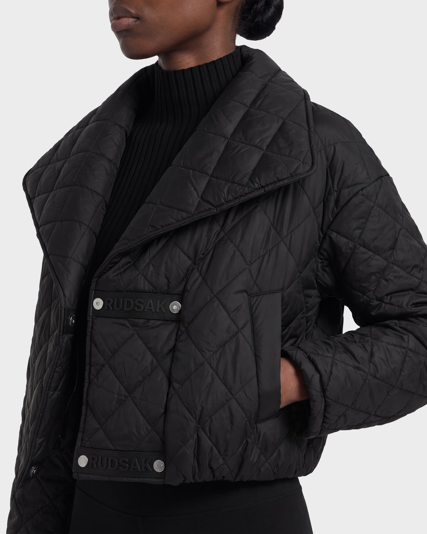 A person wearing the Rudsak KALEE - 8124005 Black quilted jacket featuring large lapels, snap buttons, and prominent Rudsak branding, layered over a ribbed black turtleneck sweater. This women's light quilted jacket brings a modern edge to a classic silhouette.