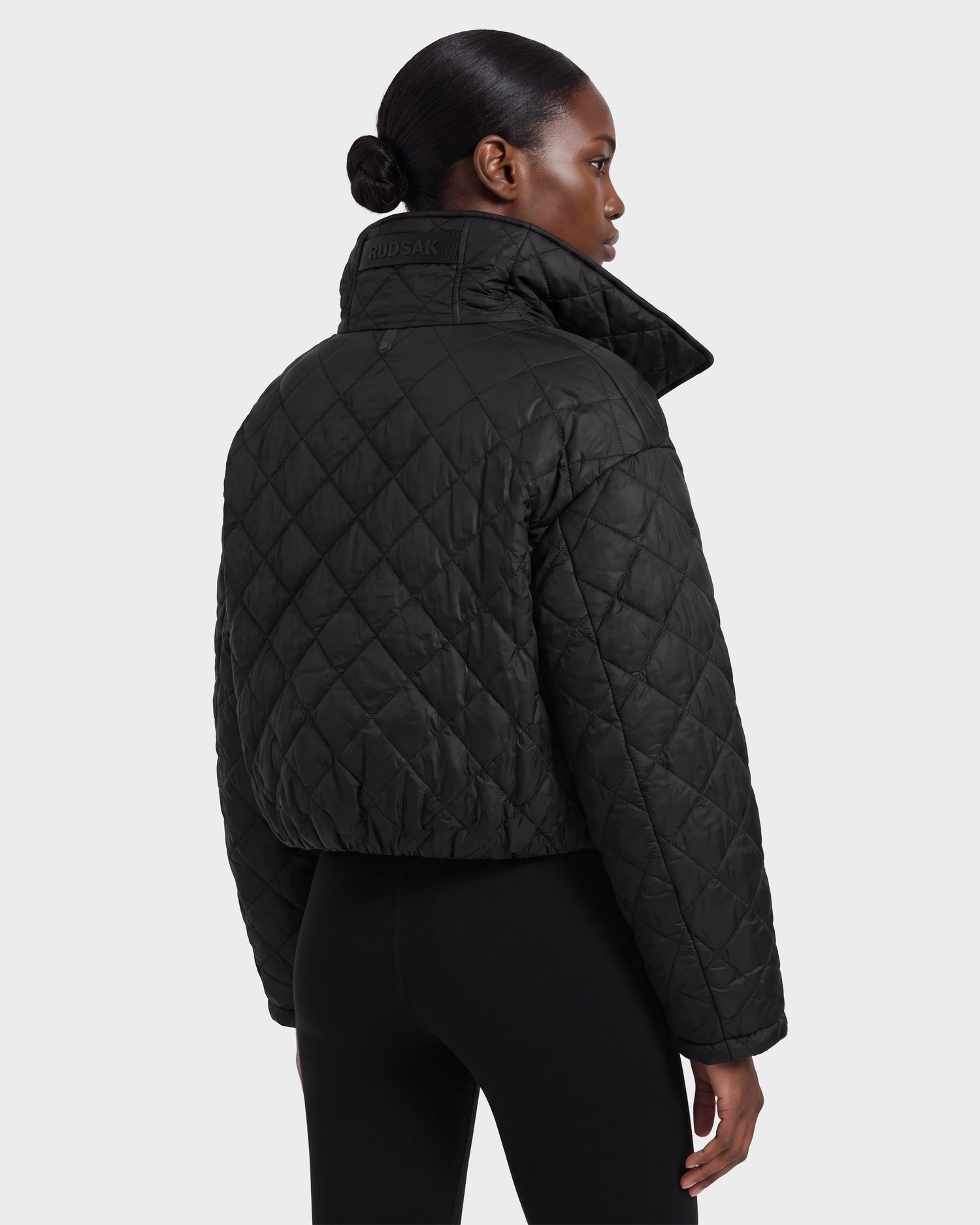 A person is wearing the Rudsak KALEE - 8124005 Black women’s quilted light jacket, featuring a cropped silhouette and high collar, paired with black leggings. She is facing away from the camera against a plain light background. This stylish piece combines warmth with modern flair.
