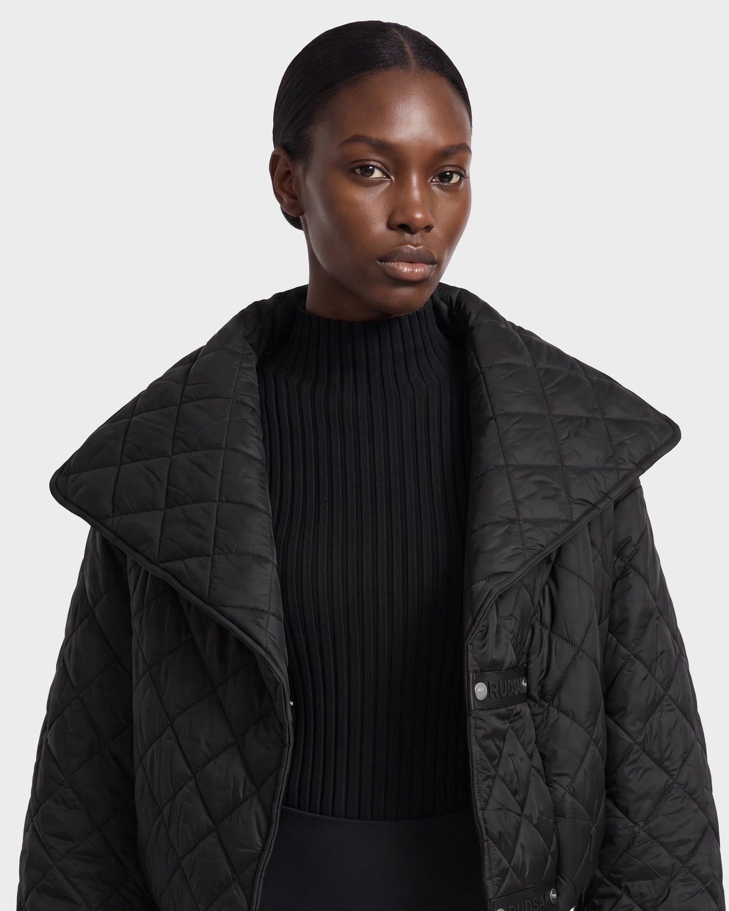 A person with smooth, dark skin and short, pulled-back hair wears the Rudsak KALEE - 8124005 Black women's quilted light jacket over a ribbed black sweater, posing against a plain gray background.