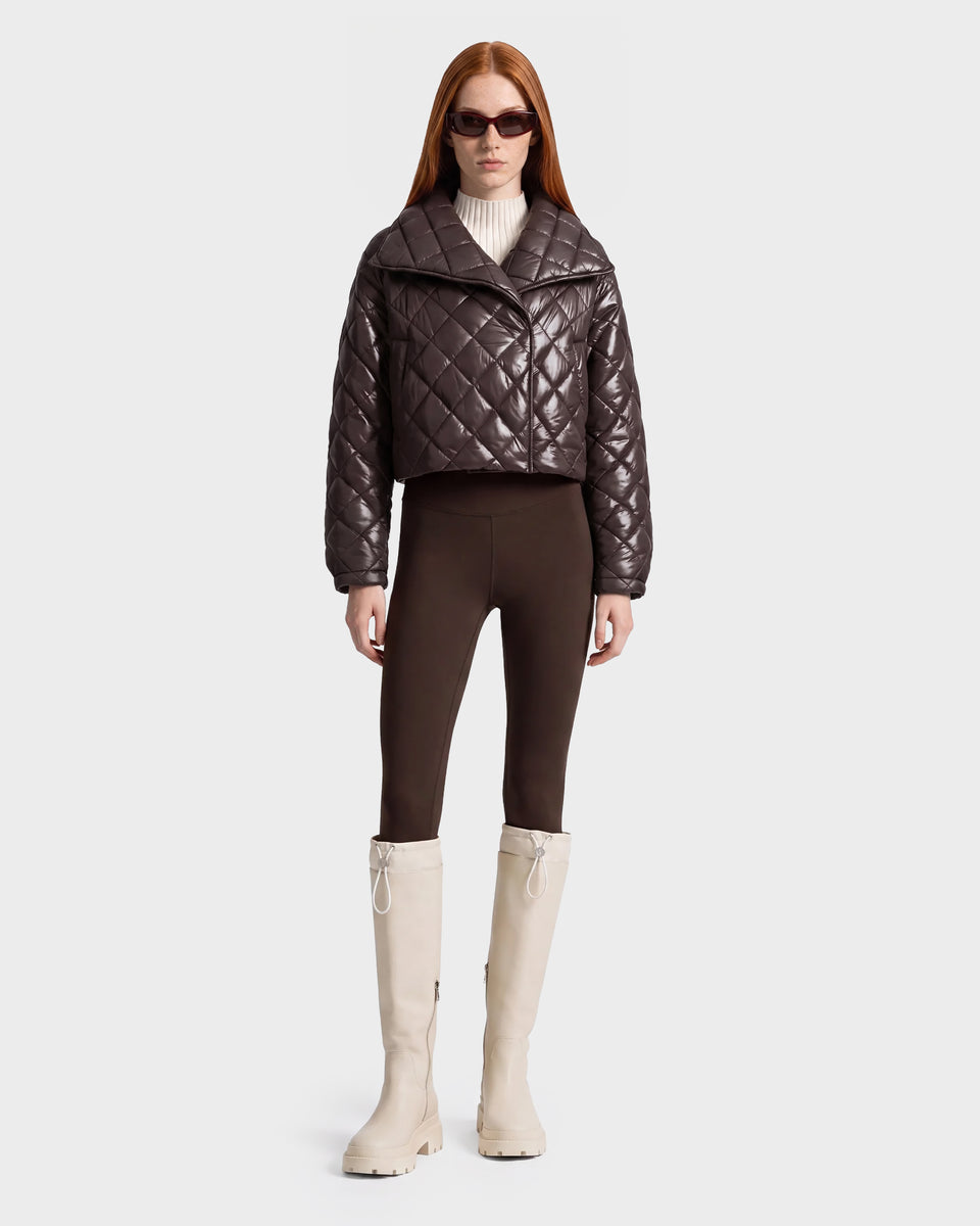 A woman stands facing forward against a plain white background, wearing the Rudsak KALEE - 8124005 Espresso women's cropped quilted jacket for a touch of active versatility, paired with brown leggings, light beige knee-high boots, stylish sunglasses, and a white turtleneck sweater.