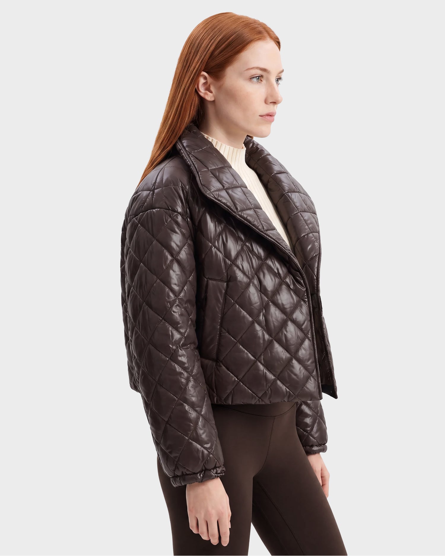 A woman with long red hair wears the Rudsak KALEE - 8124005 Espresso shiny, dark brown women's cropped quilted jacket over a cream sweater and brown leggings, standing against a plain light background.