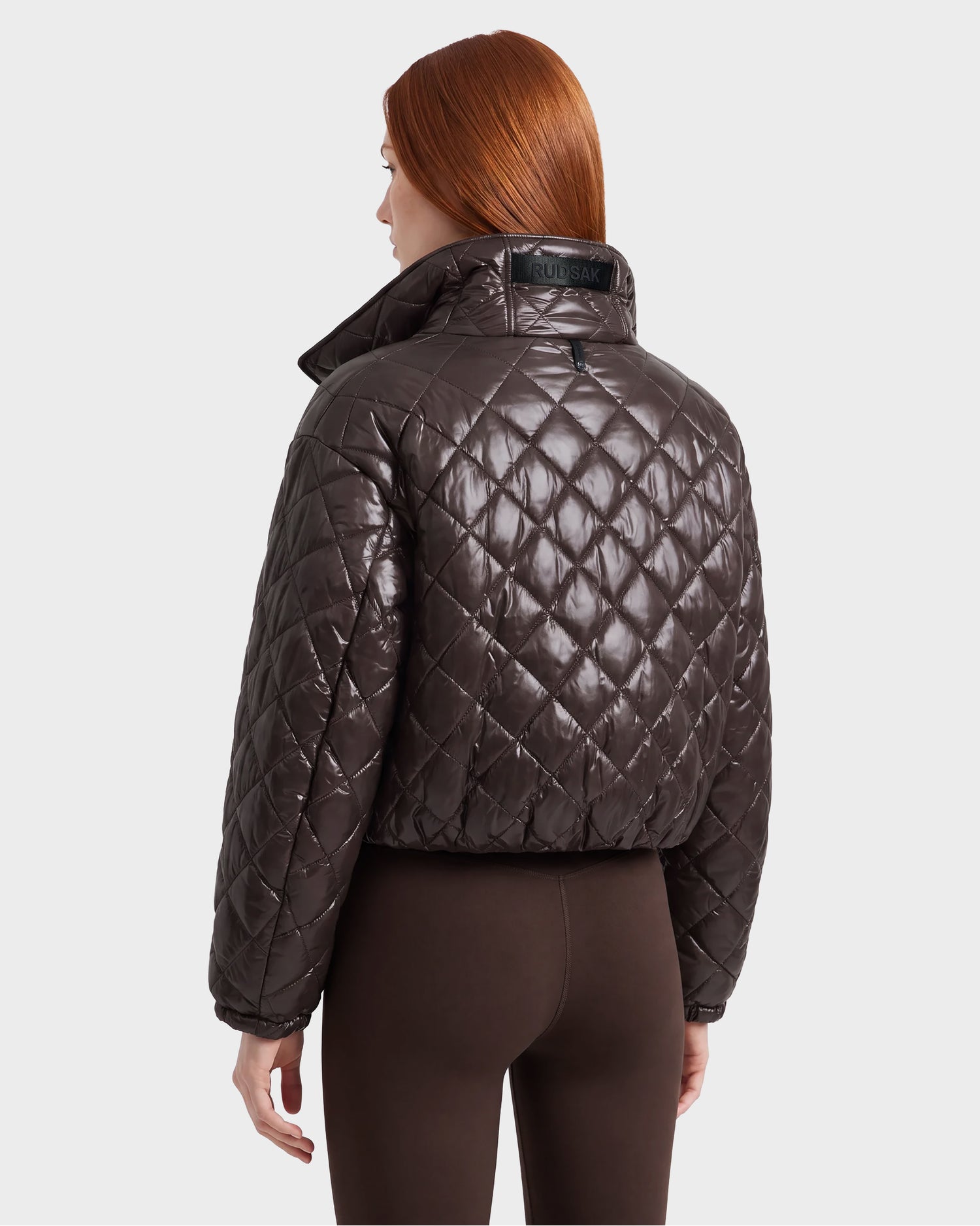 A person with long red hair is shown from the back against a plain light background, wearing the Rudsak KALEE - 8124005 Espresso women’s cropped quilted jacket and brown pants.