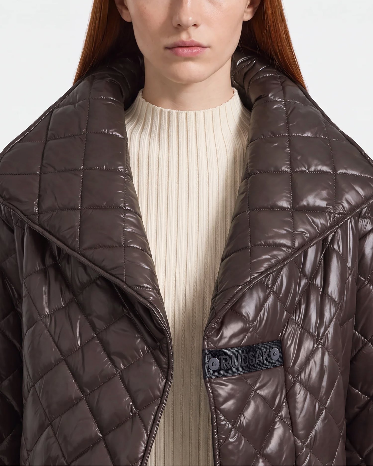 A woman with straight red hair is shown from the shoulders up wearing a cream ribbed sweater and the Rudsak KALEE - 8124005 Espresso, a shiny brown quilted light jacket with a large collar. The “Rudsak” logo patch is visible on the jacket, highlighting its active versatility.