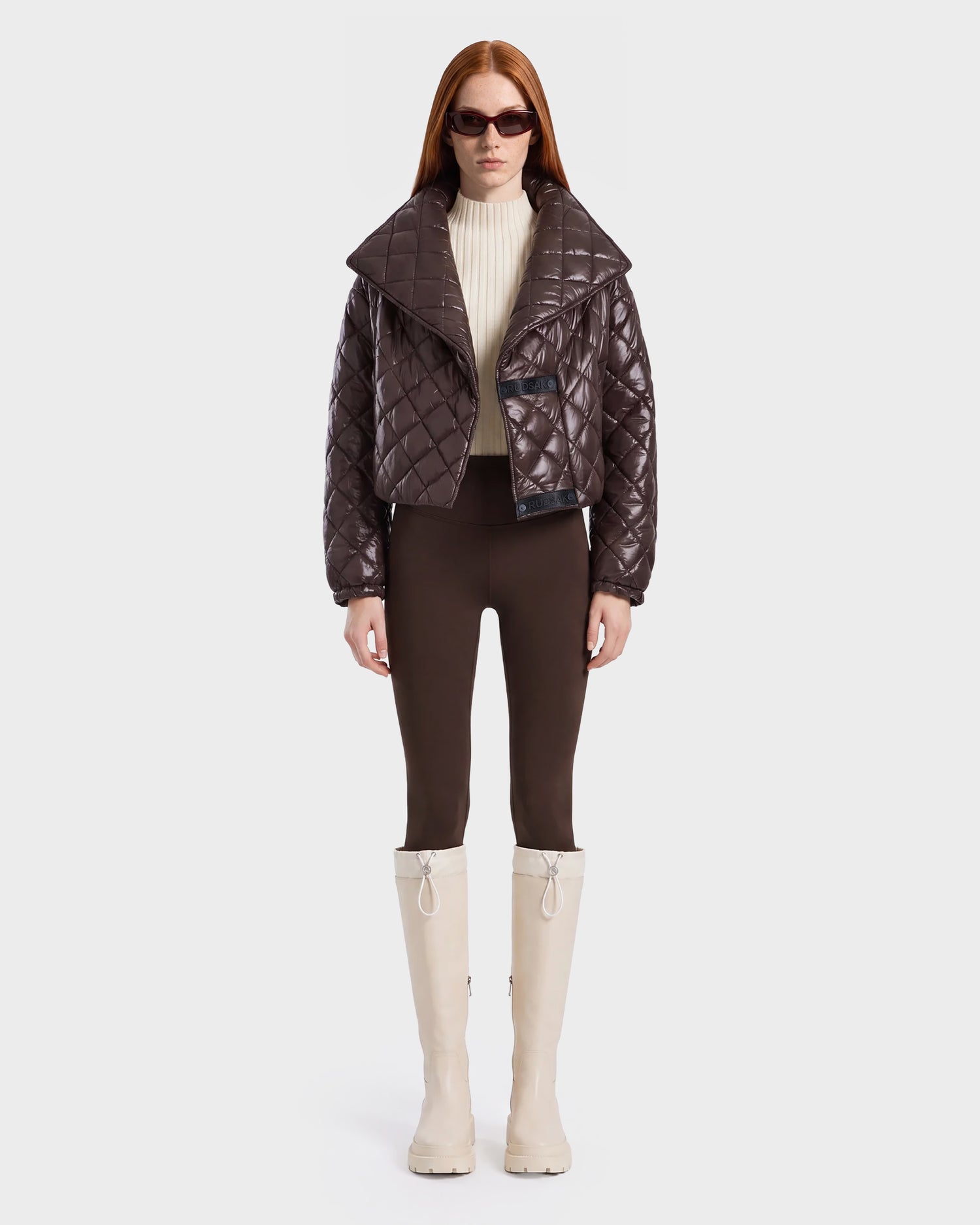 A woman stands facing forward against a plain light background, embodying active versatility in the Rudsak KALEE - 8124005 Espresso jacket styled with a cream turtleneck, brown pants, cream knee-high boots, and sunglasses for a chic and functional look.